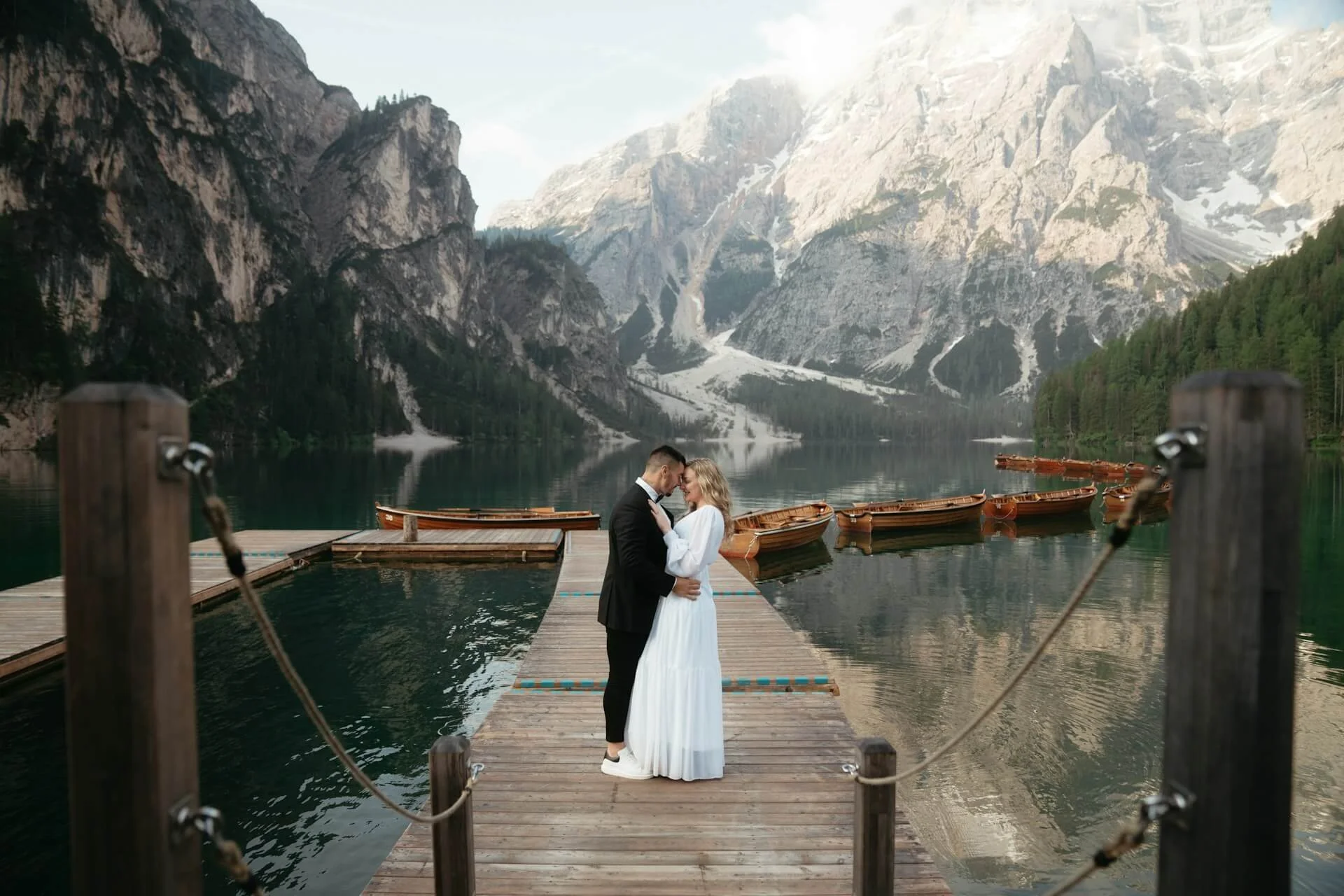 A couple dressed in wedding attire, standing on a wooden dock on a lake surrounded by mountains and evergreen trees, with boats moored nearby.