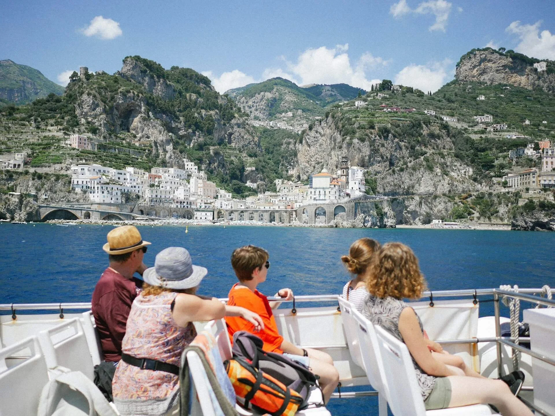 People sitting on a boat on the water with a coastal town and green mountainous landscape in the background.