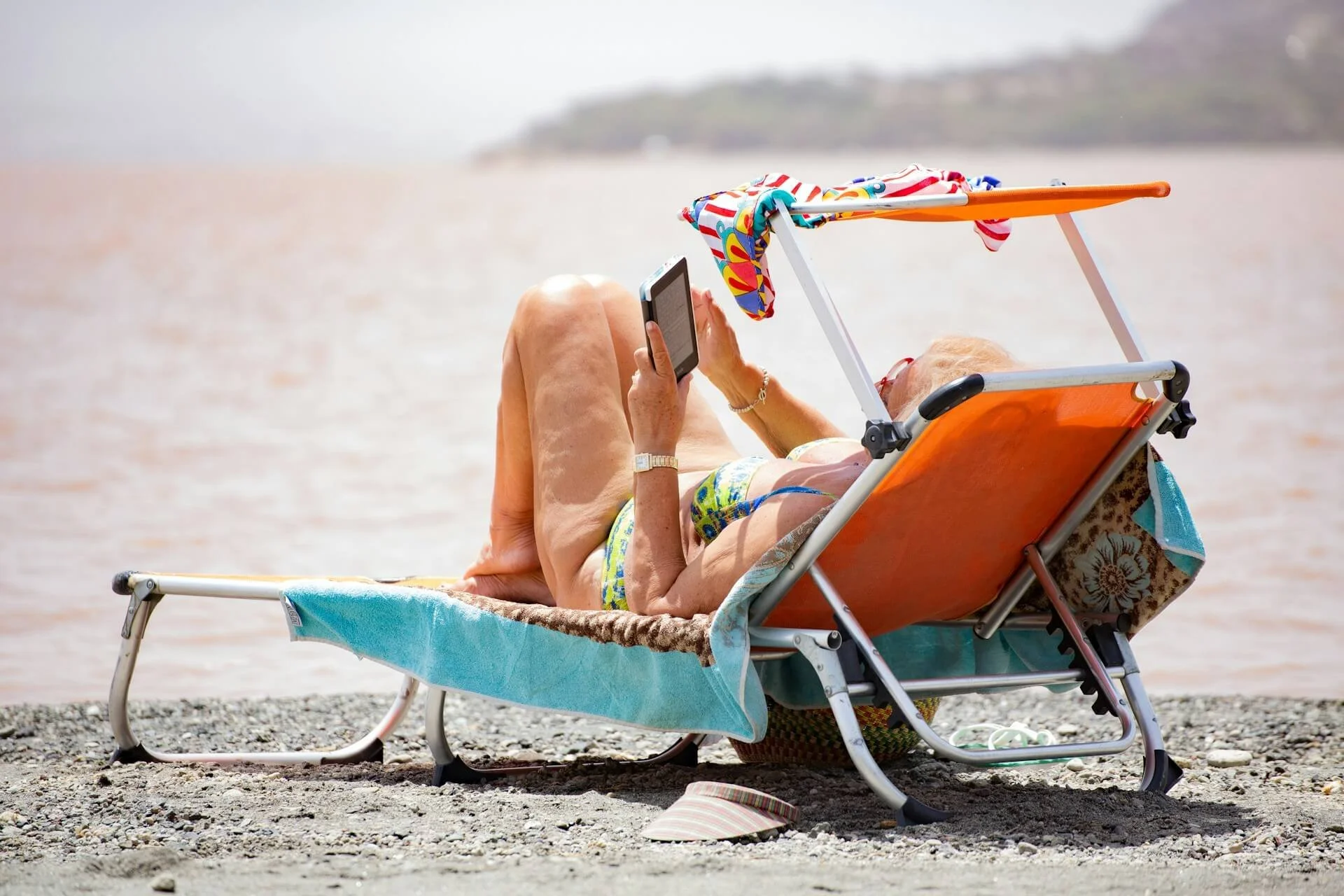 An elderly woman relaxing on a lounge chair by the water, using her phone, with a colorful towel and a hat on the rocky beach.