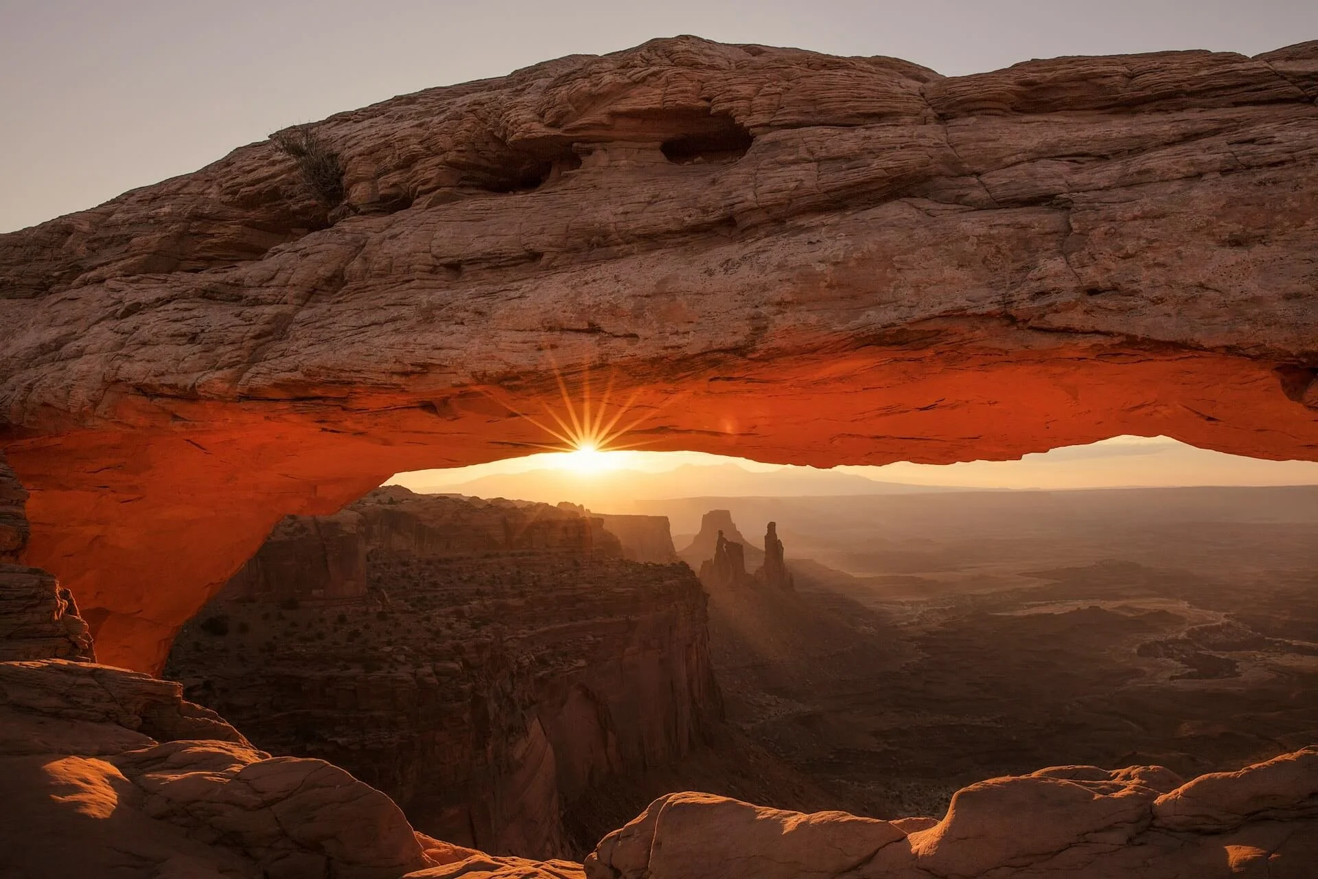 Desert landscape with rock formation and arches at sunrise or sunset, with the sun shining through an opening in the rock.