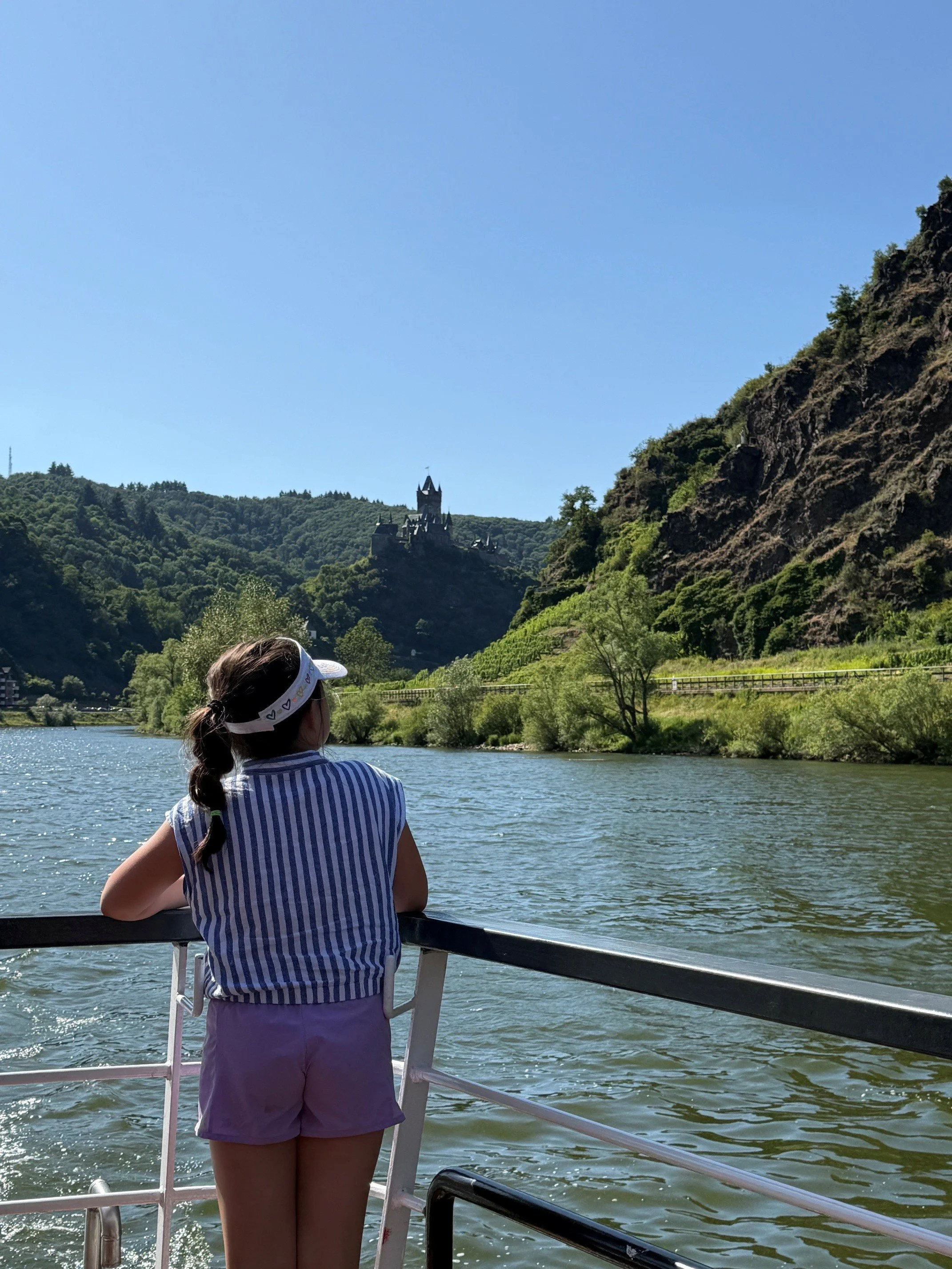 A girl with a ponytail wearing purple shorts, a striped blue and white shirt, and a white visor hat stands on a boat, looking at a castle on a hill across the river. The river has gentle ripples and lush greenery on the banks and hills in the background, with a clear blue sky.