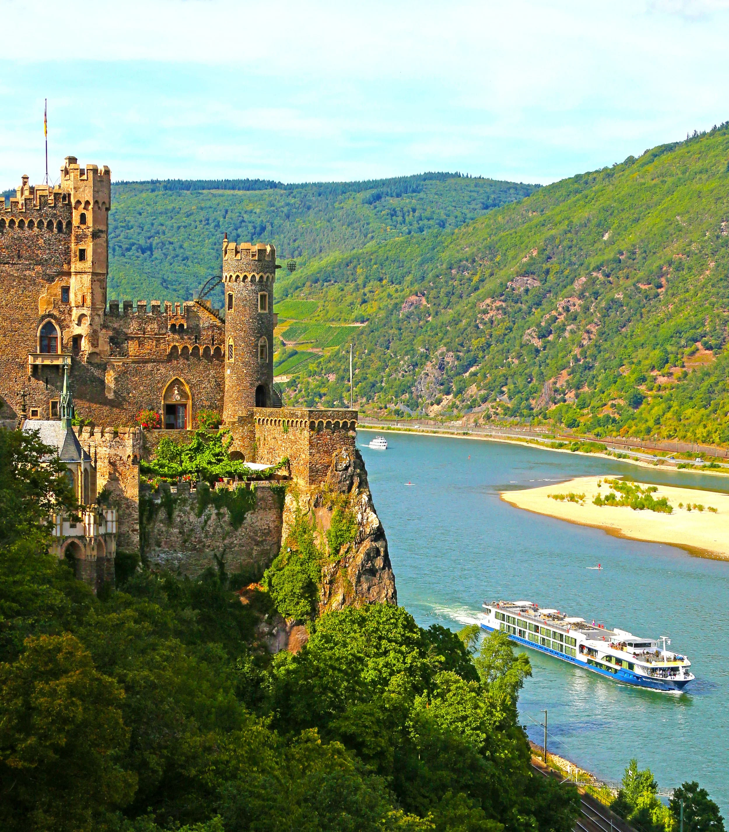 A large castle situated on a hill overlooking a river, with green forests on the hills beyond and a cruise boat sailing down the river.