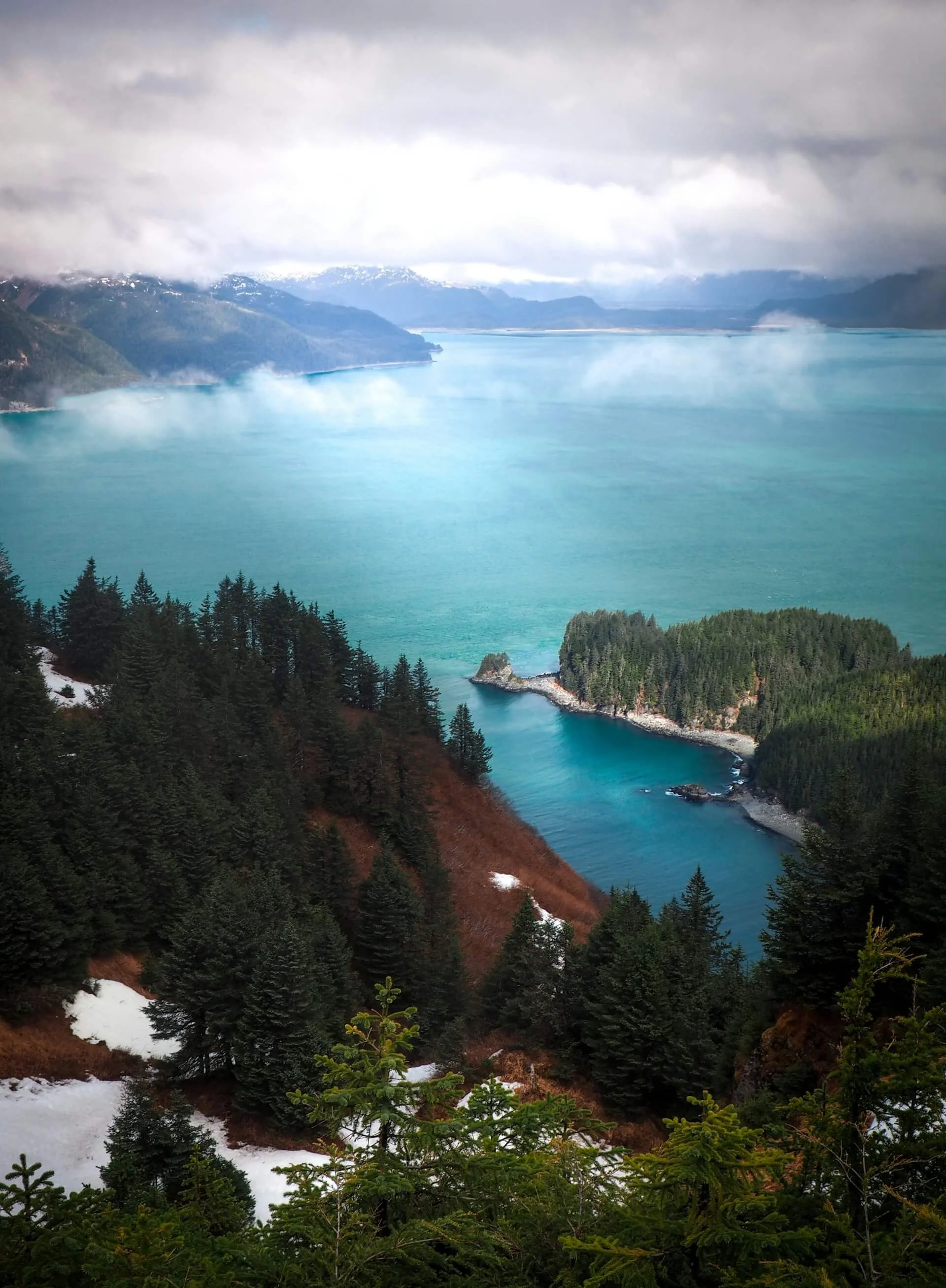 A scenic view of a large turquoise lake with surrounding forested mountains, mist, and clouds in the sky.
