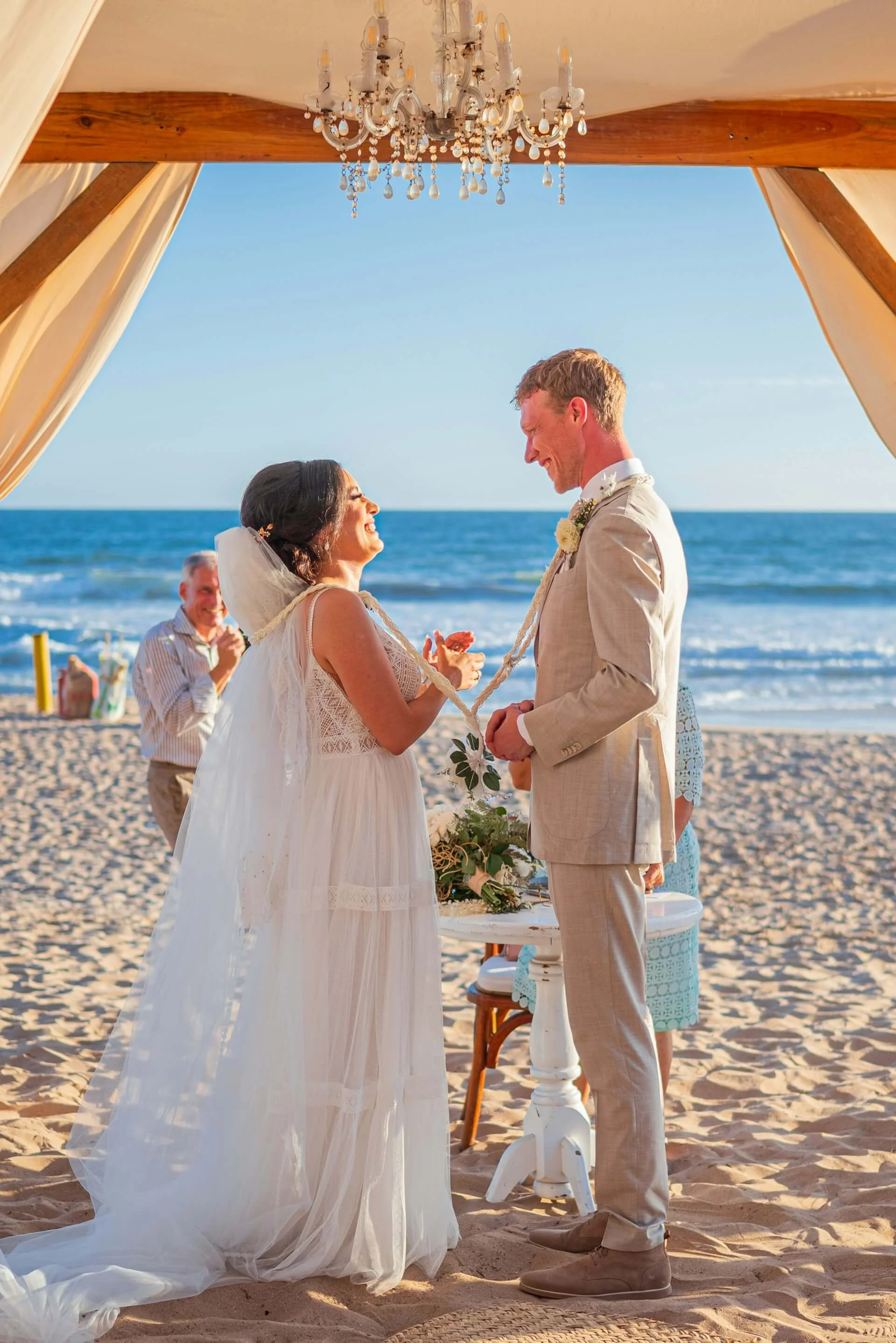 A bride and groom exchanging wedding vows at a beach wedding ceremony with the ocean in the background, under a canopy with a chandelier hanging above.