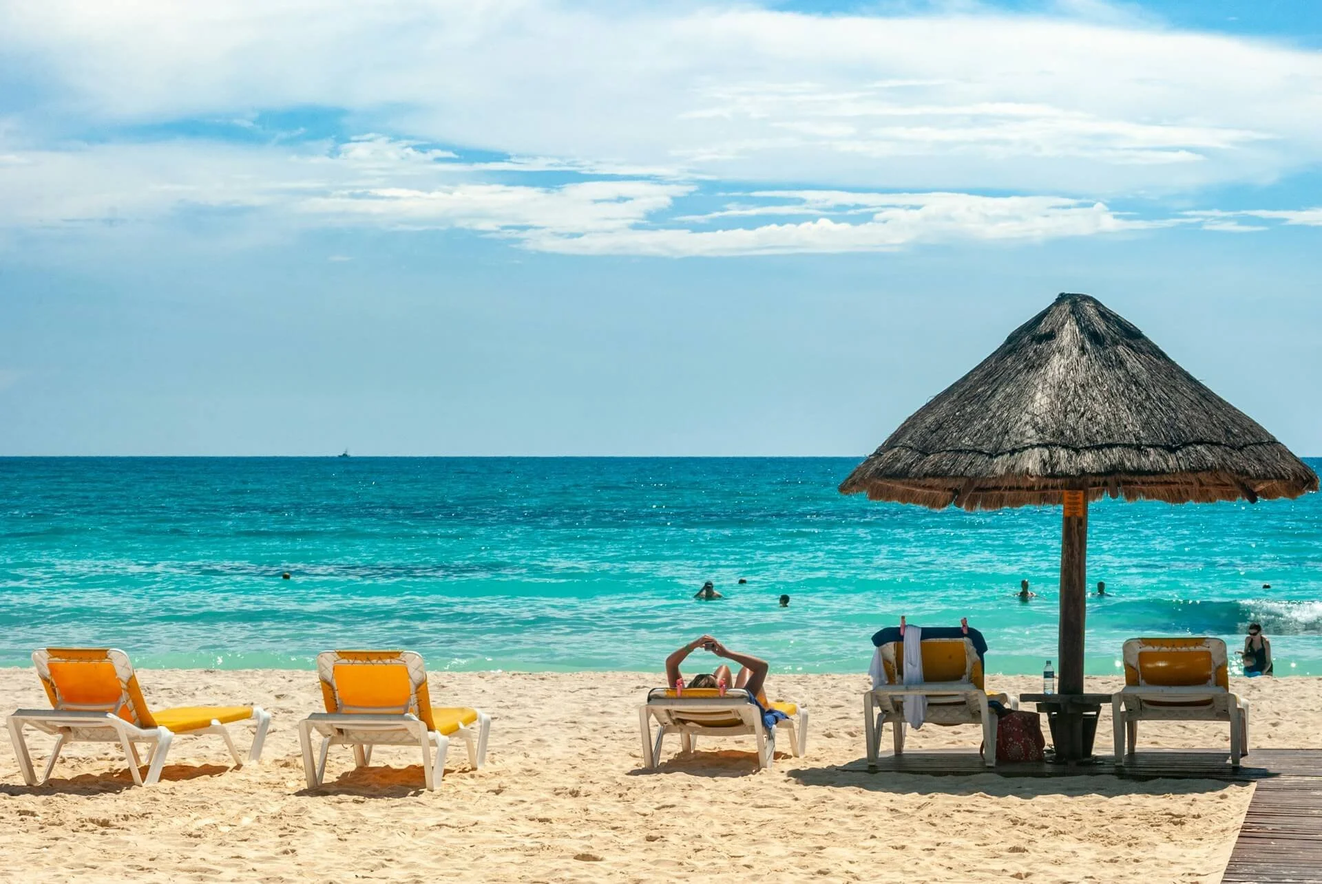 Beach with yellow lounge chairs, a straw umbrella, people in the water, and a wooden boardwalk leading to the sandy shore, ocean, and cloudy sky.