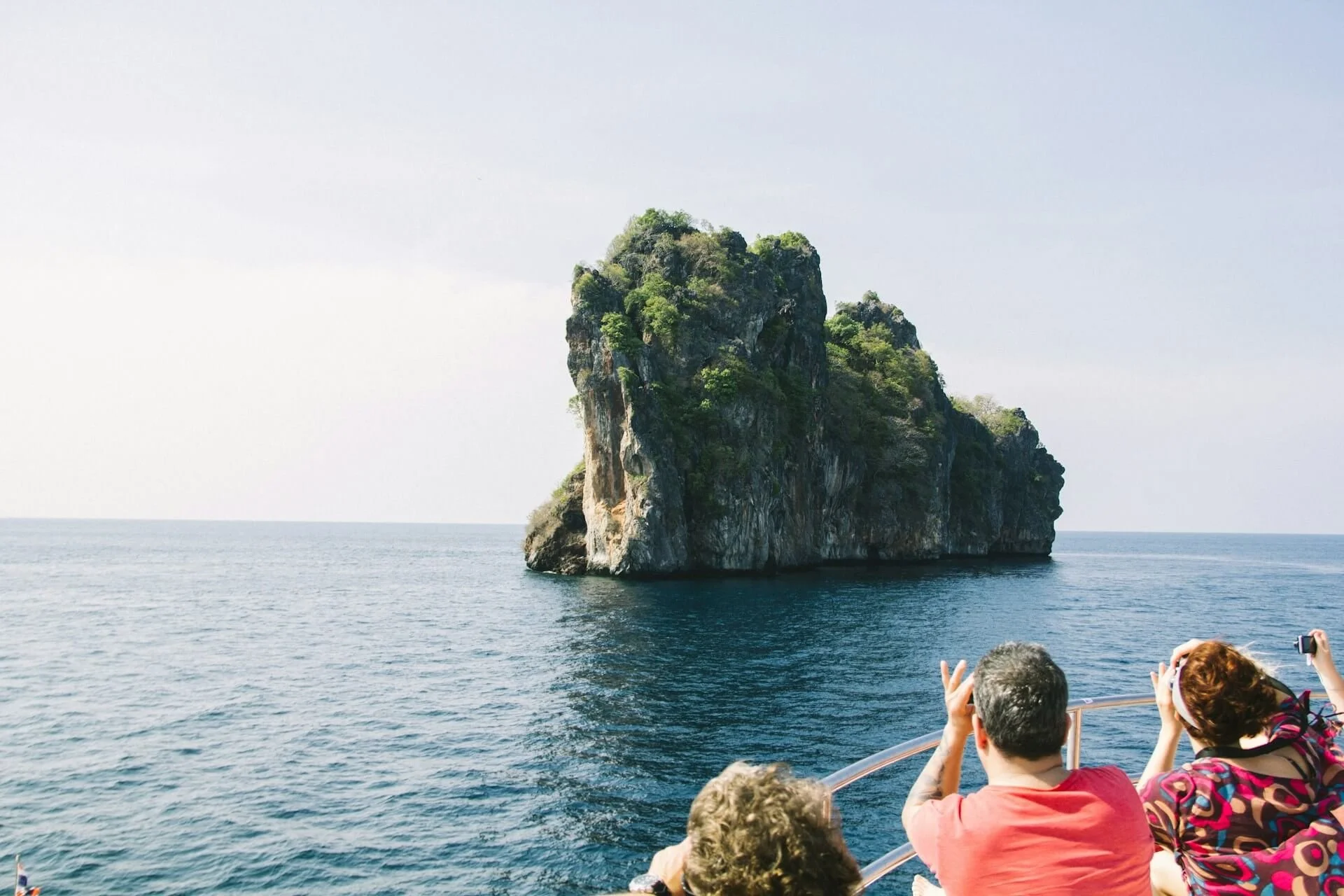 Tourists on a boat taking photos of a large rock formation with greenery rising from water in the ocean