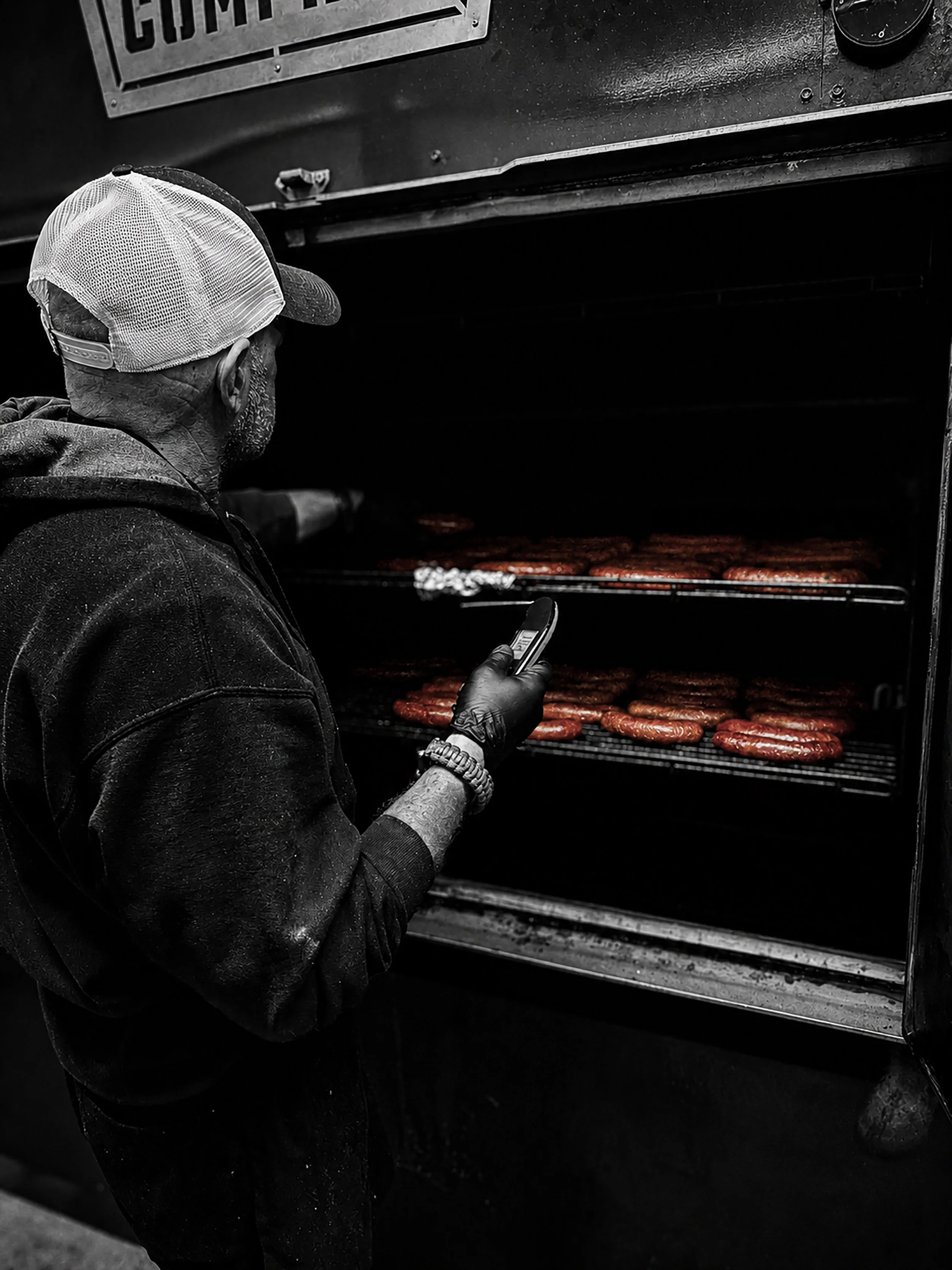 A man wearing a cap and gloves is looking at a barbecue grill with sausages cooking inside, while holding a phone in his right hand.