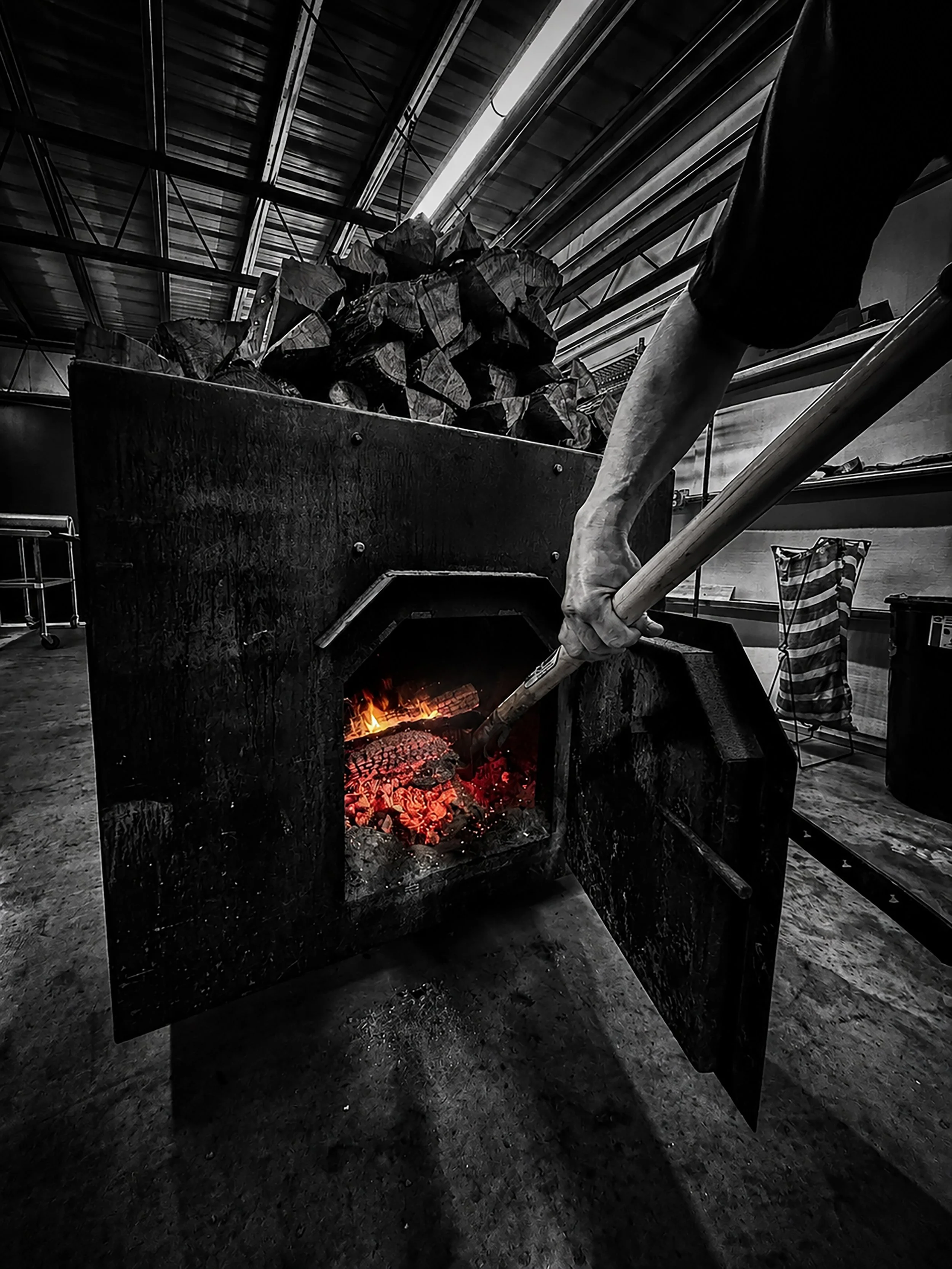 Black and white photo of a fireplace with a small fire burning inside. To the right, a large human arm is holding a long metal poker. In the background, there are stacked logs of wood on a raised platform. Various objects, including a striped bag, are visible around the space.
