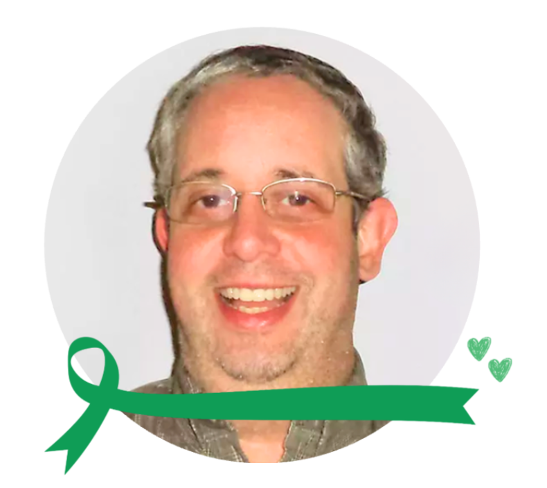 A smiling man with glasses and a beard, wearing a gray shirt, surrounded by a white circular background with green ribbon and small green hearts.