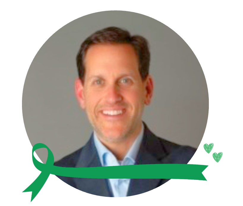 A smiling man with short brown hair, wearing a dark blazer and light blue shirt, in a gray circle background with a green ribbon and two green hearts at the bottom right.