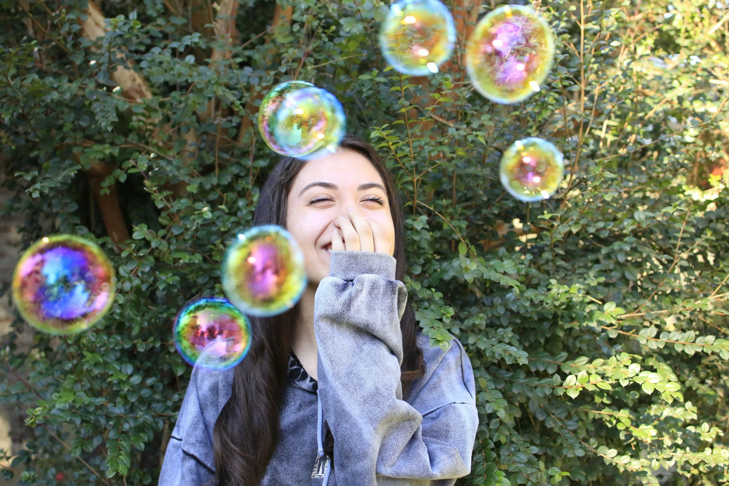 A young woman laughing and covering her mouth with her hand stands in front of green bushes, with soap bubbles floating around her.