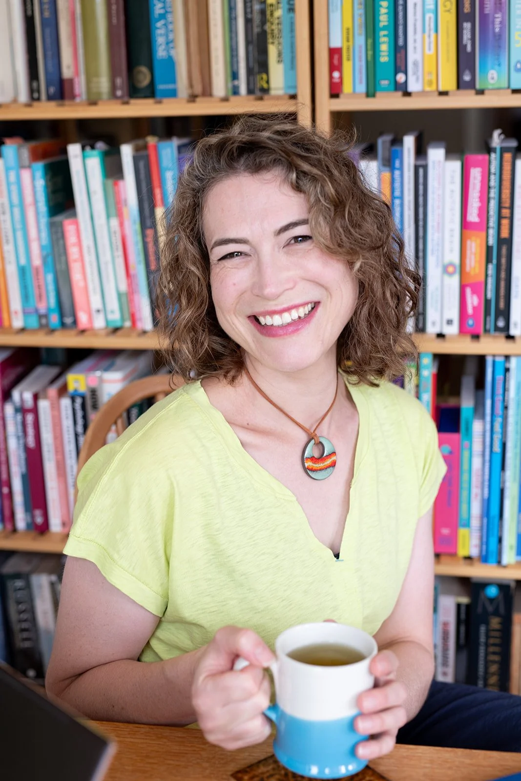 A smiling woman with curly brown hair, wearing a yellow shirt, holding a mug of tea, sitting in front of a bookshelf filled with colorful books.