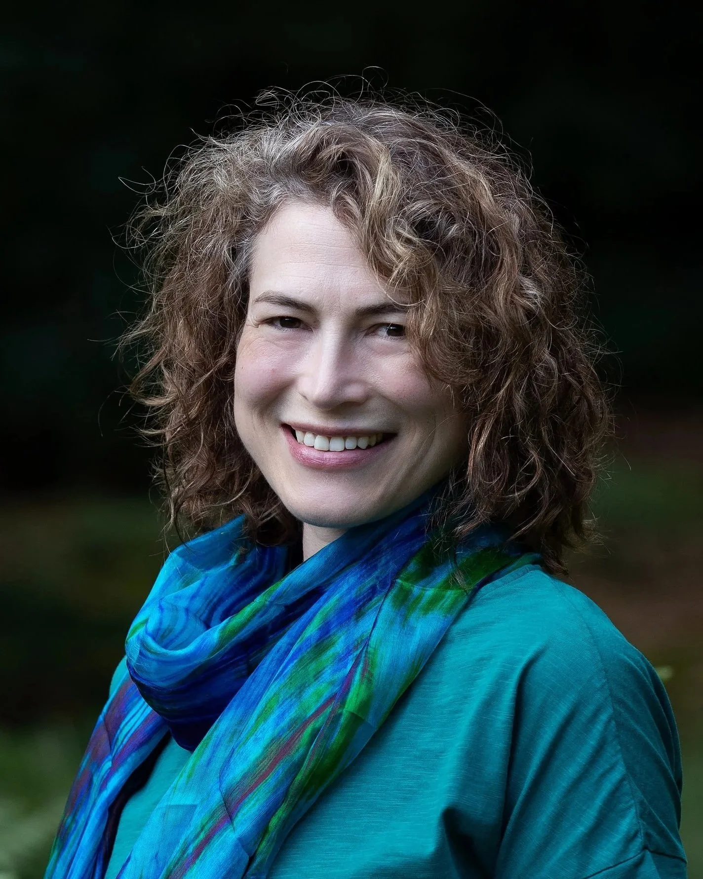 A woman with curly brown hair smiling outdoors, wearing a teal top and a blue-green scarf.