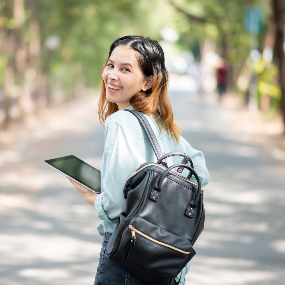 Young woman with long hair holding a tablet and carrying a backpack, standing on a tree-lined street with sunlight filtering through the leaves.