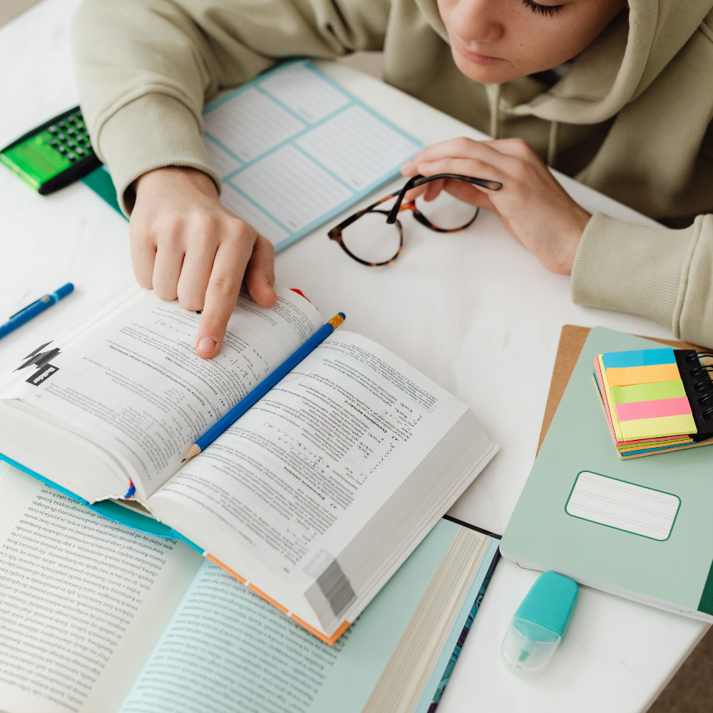 A tenaged boy studying with an open book on a desk, pointing at a page with a blue pencil, with glasses, a notebook with colorful sticky notes, a highlighter, and other school supplies around.