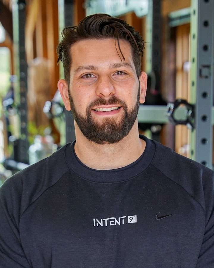 A man with dark hair, beard, and mustache smiling at the camera, wearing a black T-shirt labeled 'INTENT 91' in a gym with wooden walls and workout equipment in the background.
