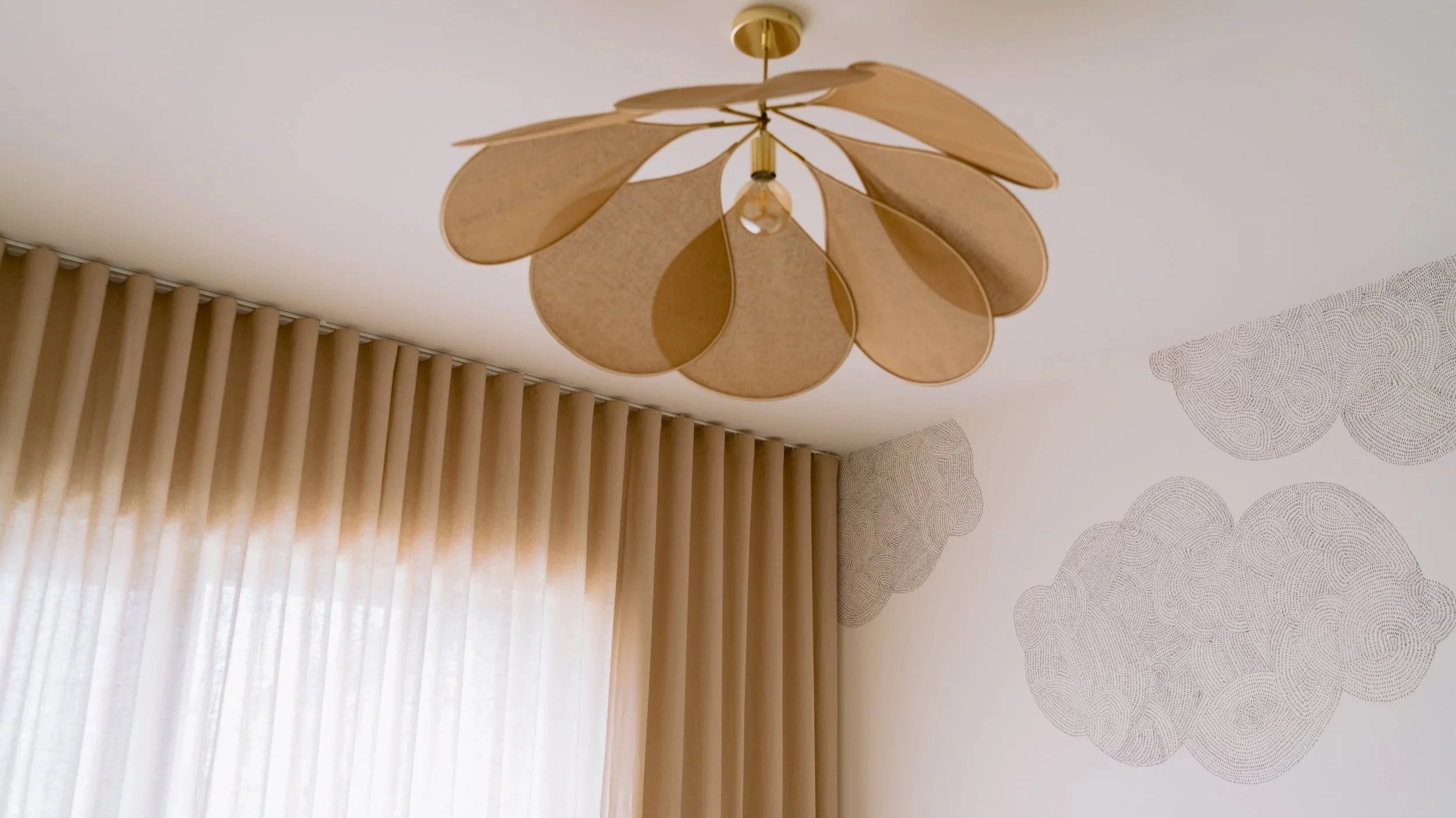 Ceiling light fixture resembling a flower with petal-shaped wooden panels, in a room with beige curtains and cloud-shaped wall decorations.