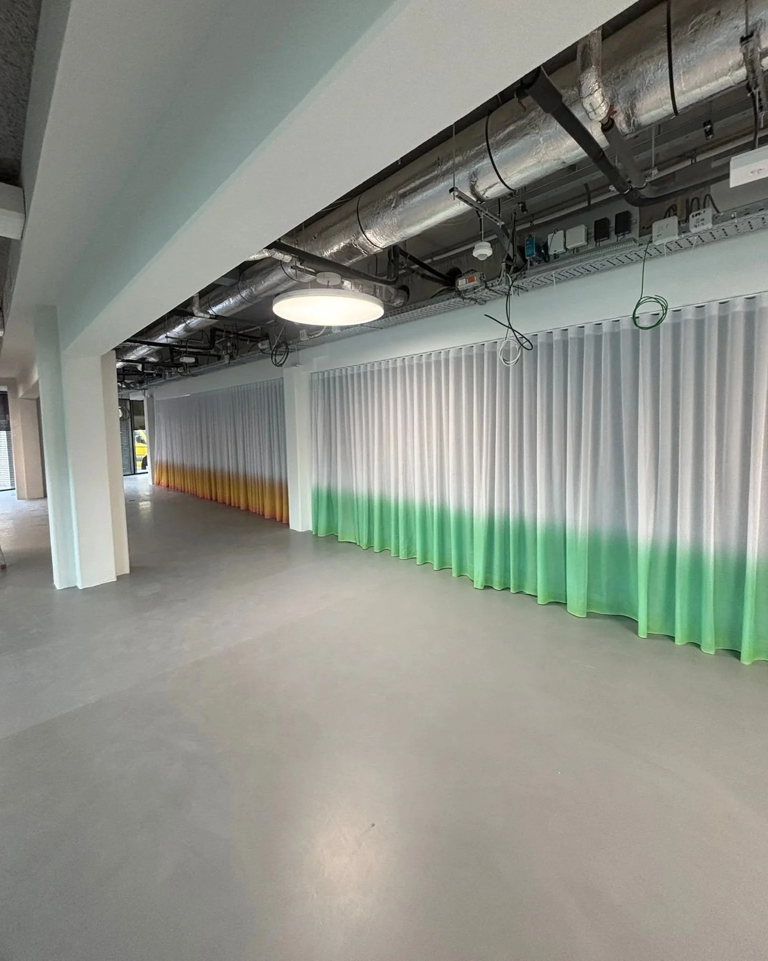 Interior of an unfinished commercial space with exposed ceiling pipes and electrical wiring, a white wall with a gradient curtain starting yellow at the top and fading to green at the bottom, and a large overhead light.