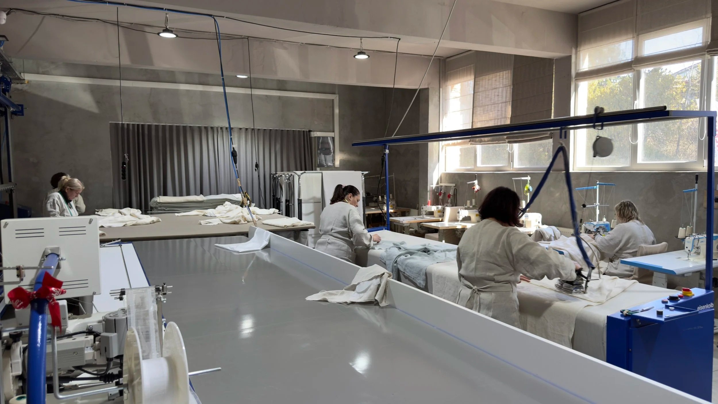 Workers in white lab coats sewing fabric on sewing machines in a bright, industrial workshop with large windows.