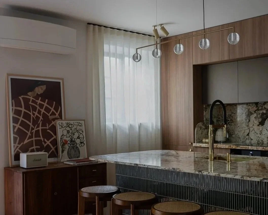 Modern kitchen with marble countertop, brass faucet, wood and gray cabinetry, decorative wall art, and pendant lights.