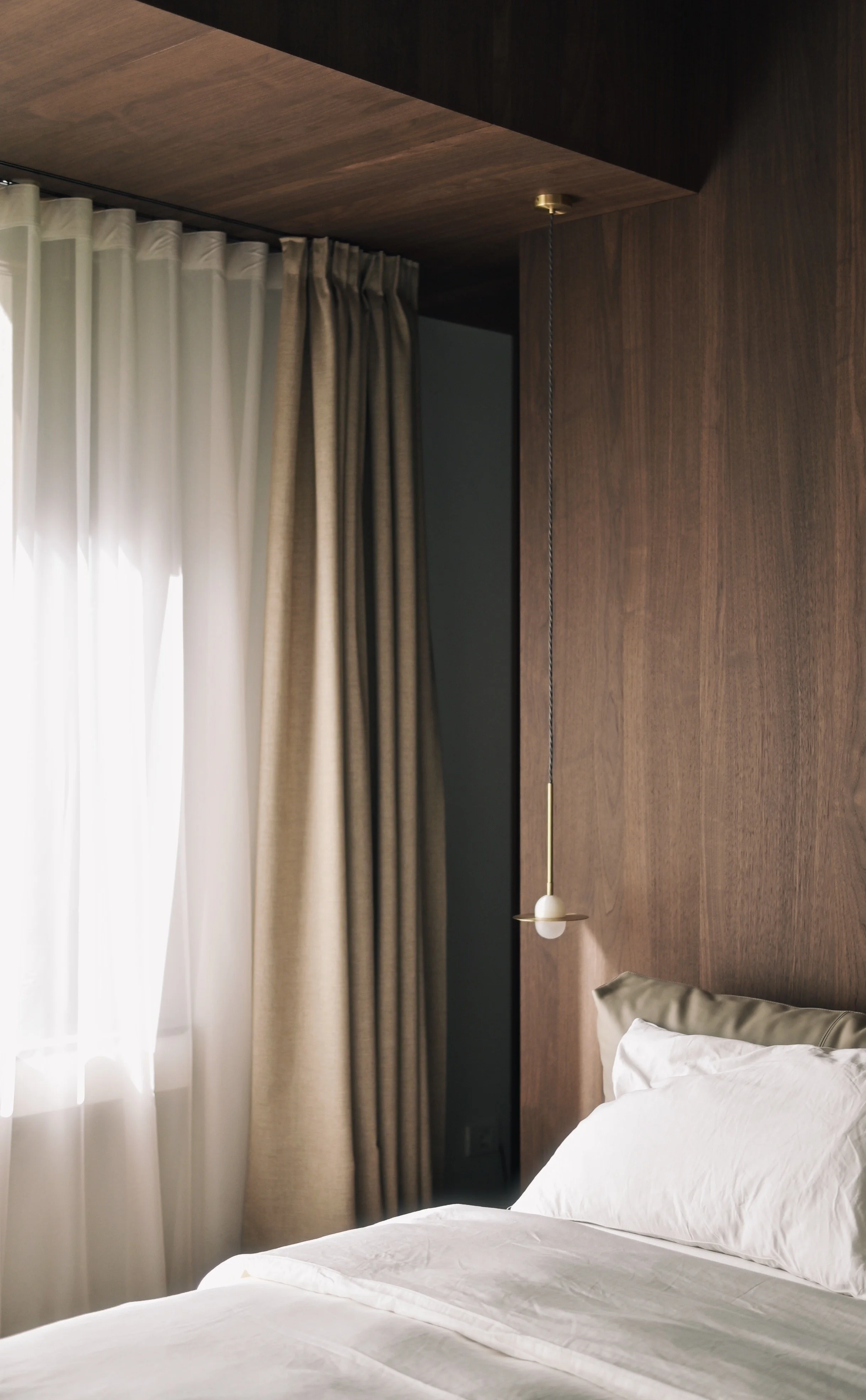 A bed with white bedding and pillows, next to a wooden headboard wall, with a hanging light and curtains covering a window.