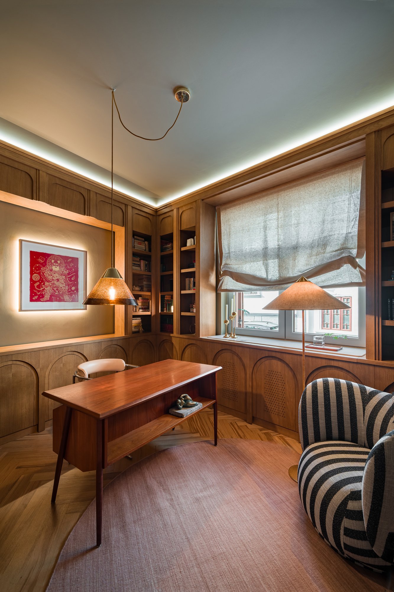 A cozy home office with wooden paneling, built-in bookshelves filled with books, a window with a beige Roman shade, a wooden desk, a striped armchair, a round pink rug, and soft lighting from hanging and table lamps.