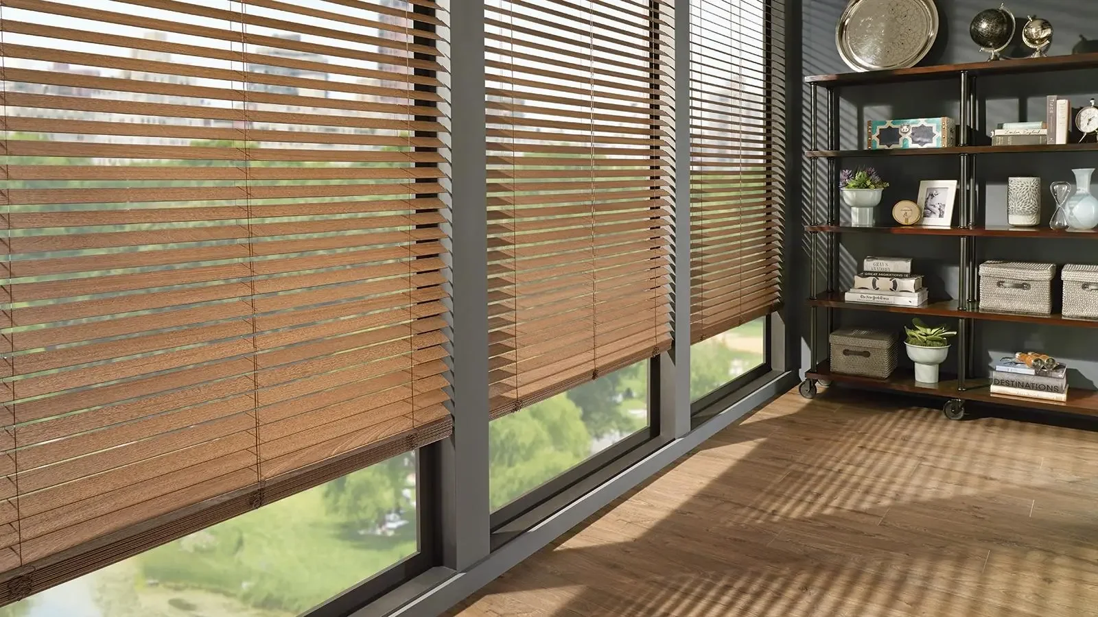 Room with windows covered by wooden blinds, a metal and wood shelving unit with decorative items, plants, books, and storage baskets, and hardwood floors.