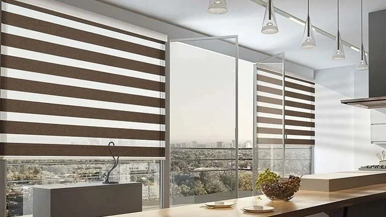 Modern kitchen with large windows covered by striped roller blinds, a view of cityscape and trees, kitchen counter with plates and a bowl of fruit, and ceiling lights.