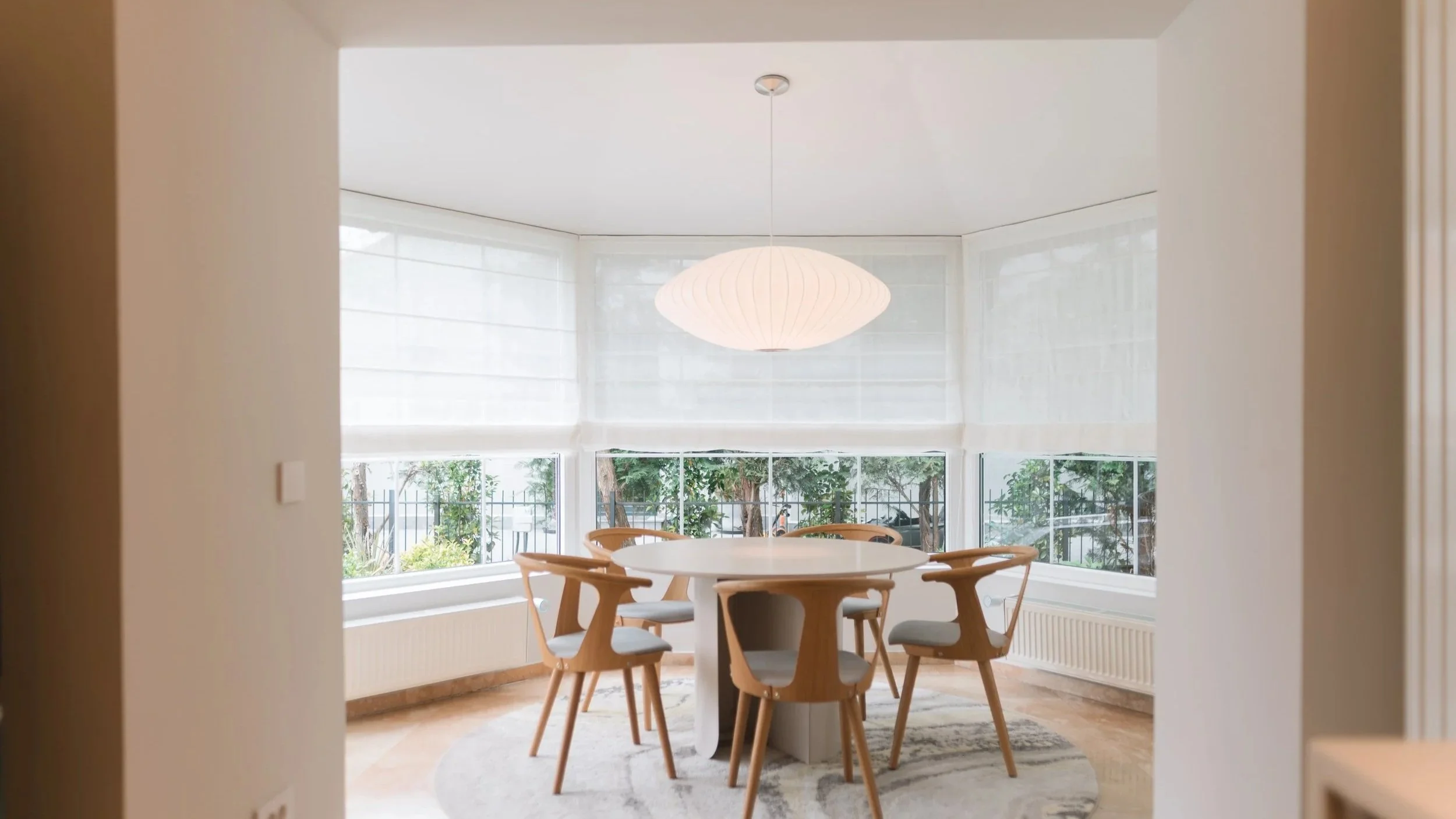 Bright dining room with a round white table, six wooden chairs with gray cushions, large windows with white blinds, a white ceiling with a round pendant light, and a light-colored rug on a wooden floor.