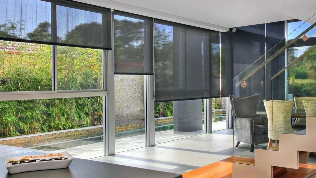Modern interior living room with large floor-to-ceiling windows, gray window shades, and a view of greenery outside; includes a gray armchair and a part of a dining table with chairs.