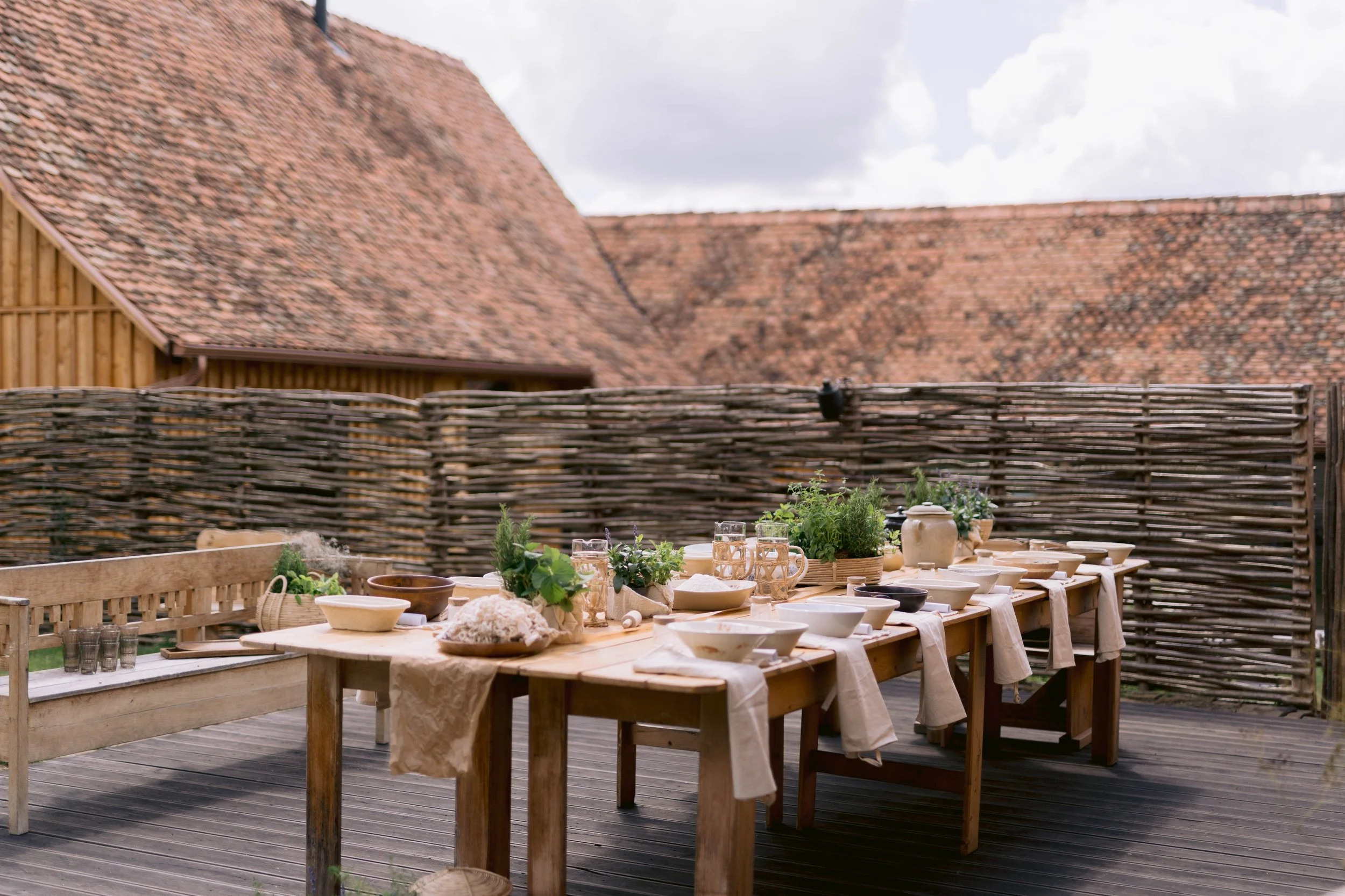 Outdoor dining table set with dishes, glasses, and plants on a wooden deck, bordered by a woven wooden fence, with rustic buildings and a cloudy sky in the background.