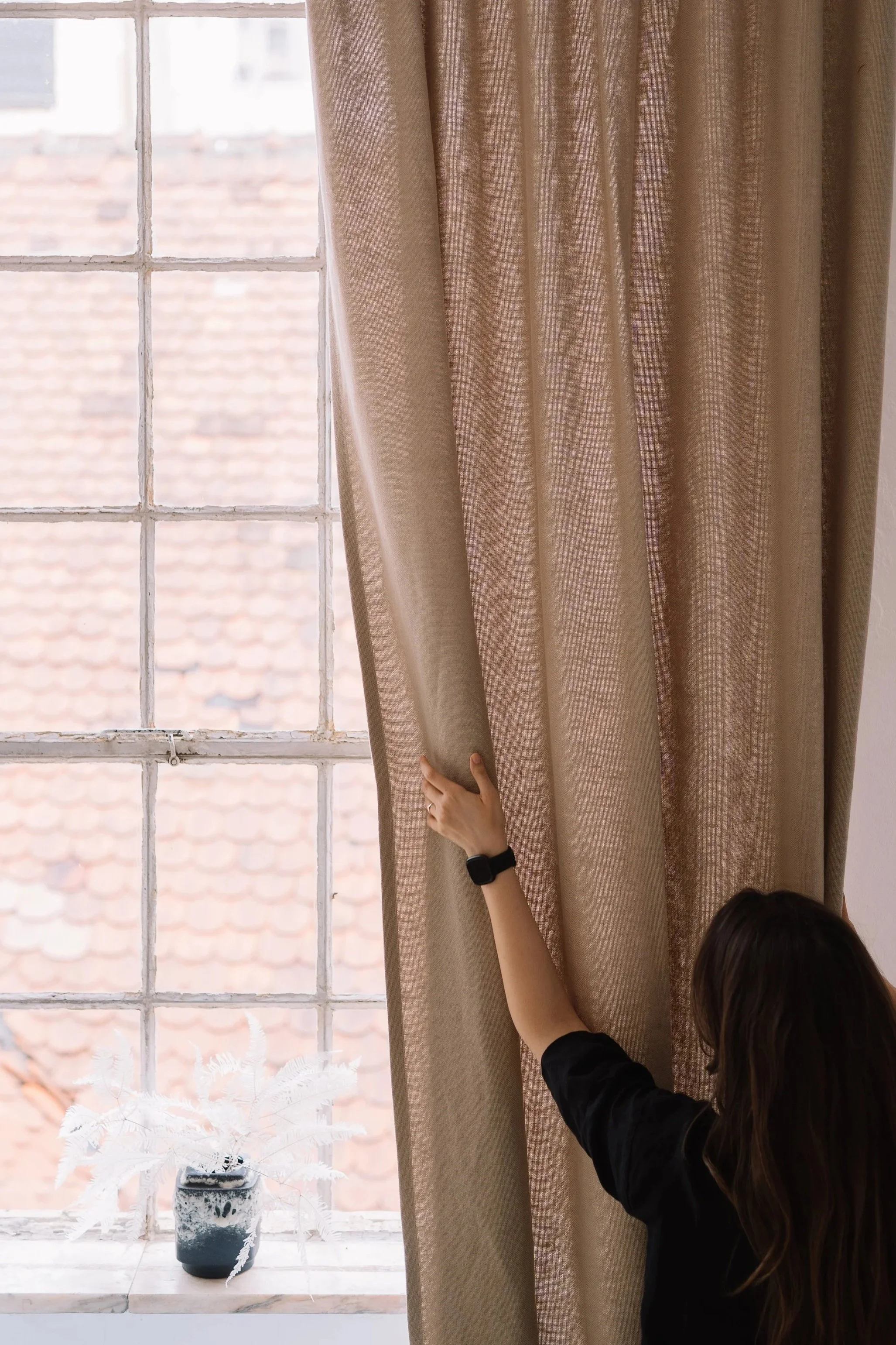 Person adjusting beige curtains by a window with a black potted plant on the windowsill.