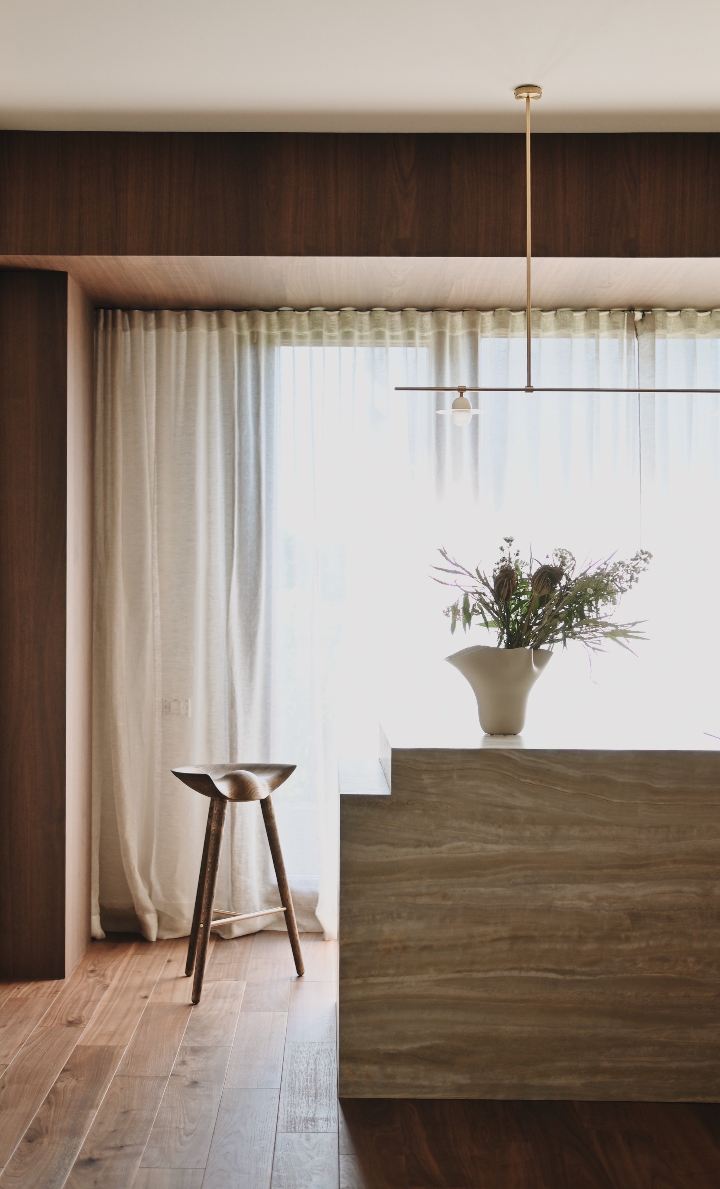 A minimalist interior with a wooden stool, a large flower vase with a bouquet, a marble countertop, and sheer white curtains allowing natural light to fill the room.