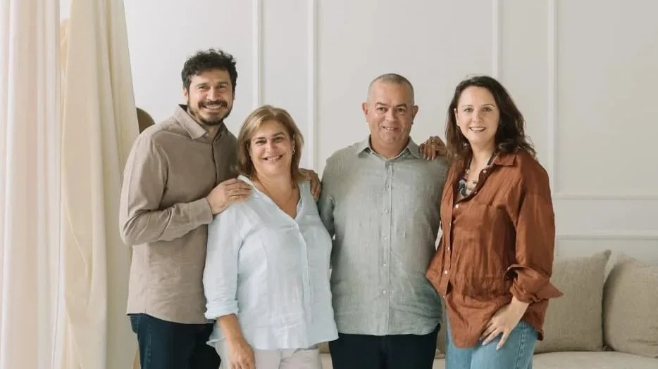 Group of four people, three women and one man, smiling and standing close together in a well-lit living room with white walls and cream curtains.