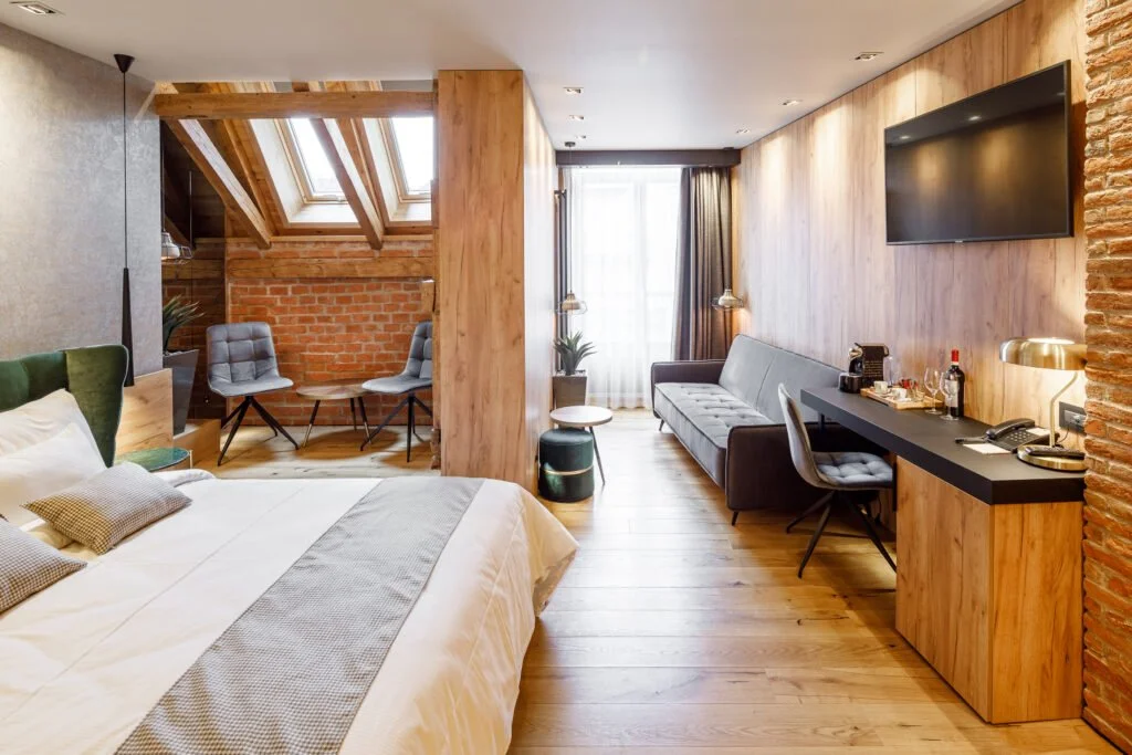 Modern hotel room with a bed, sitting area, and entertainment area featuring a flat-screen TV, wood and brick accents, and natural light.