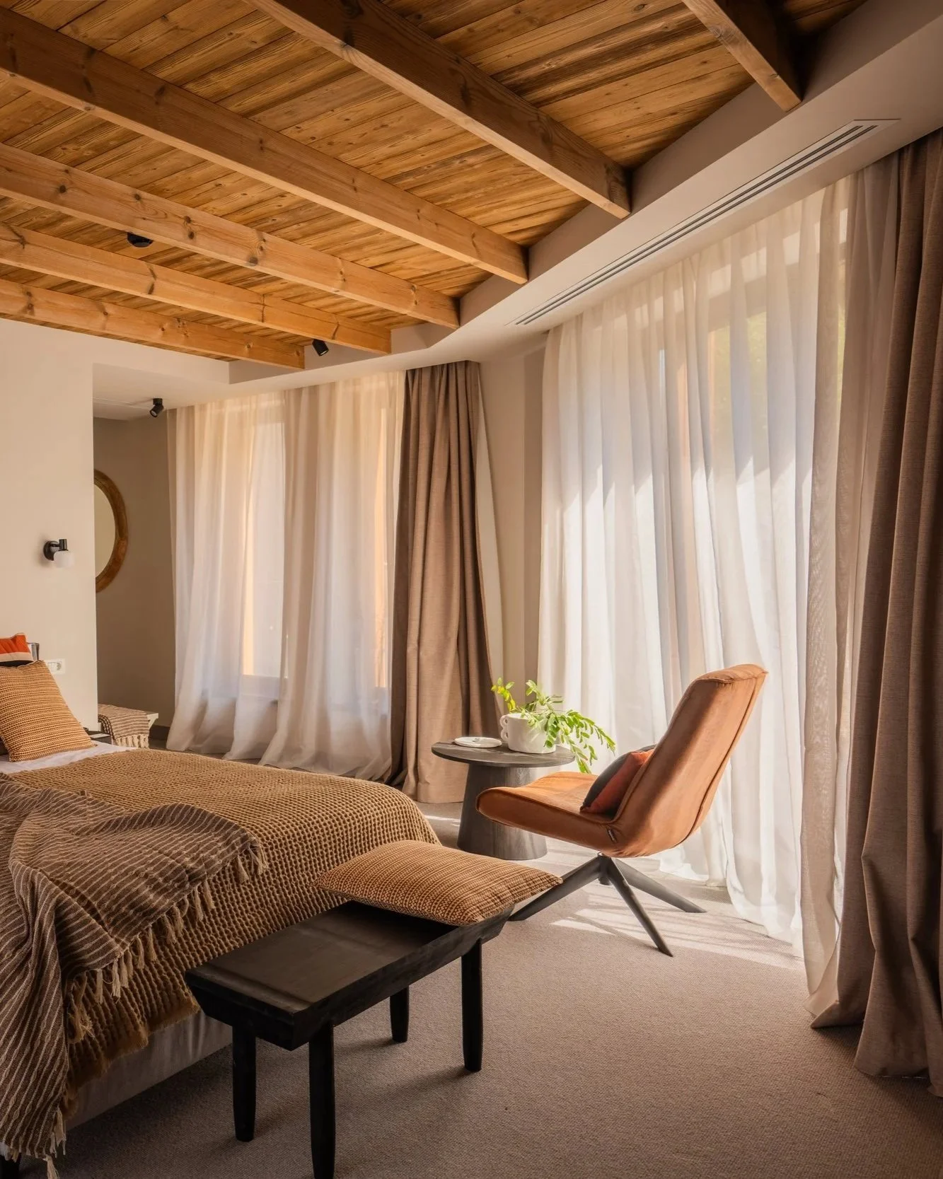 A cozy bedroom featuring a bed with brown and orange bedding, a black wooden bench at the foot of the bed, a brown upholstered chair beside a round side table with a plant, large windows with sheer white curtains and brown drapes, a wooden ceiling with exposed beams, and beige carpet flooring.