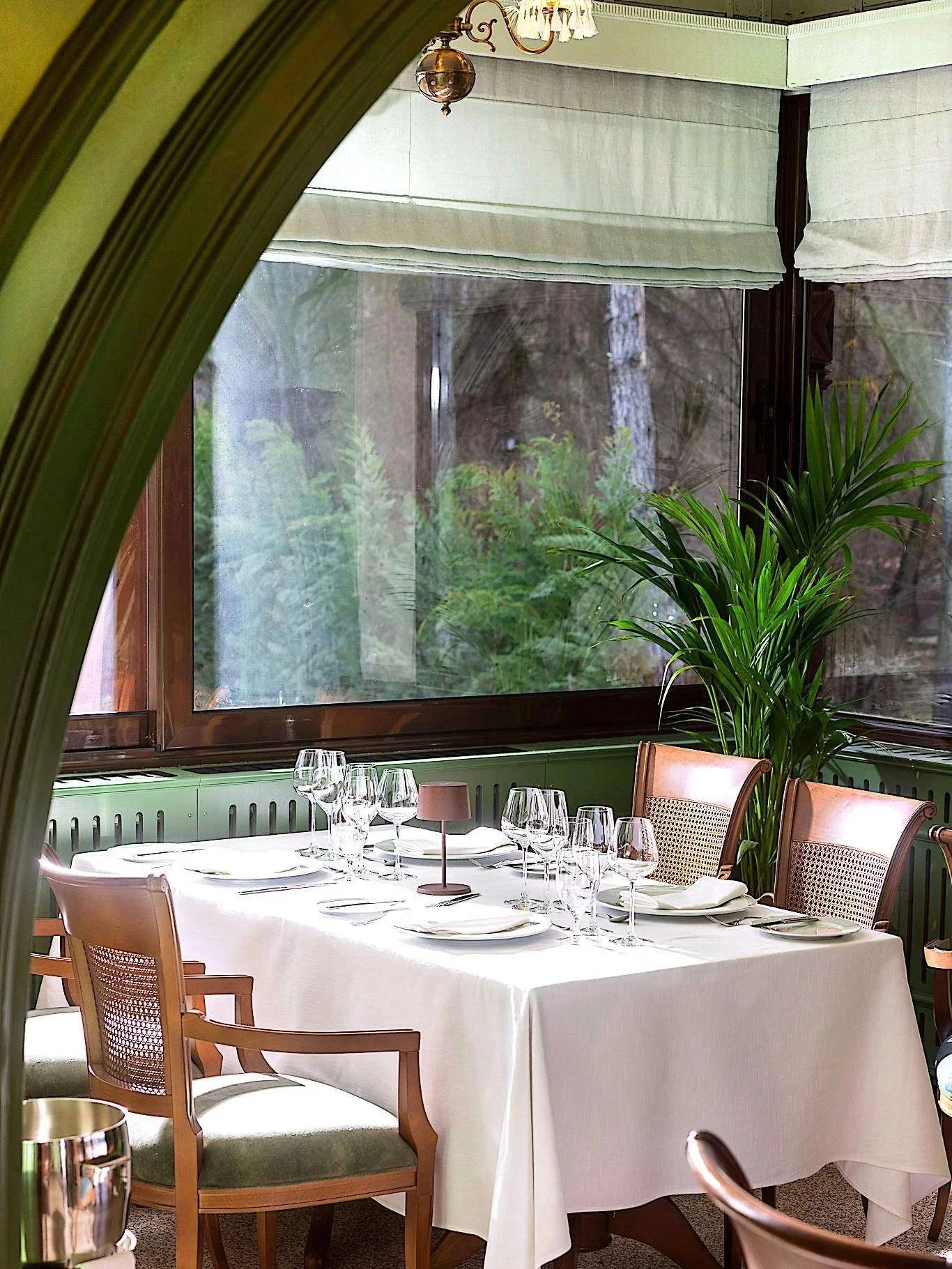 A dining table set for a meal with six wine glasses, plates, and napkins in a room with large windows, green walls, and a potted plant.
