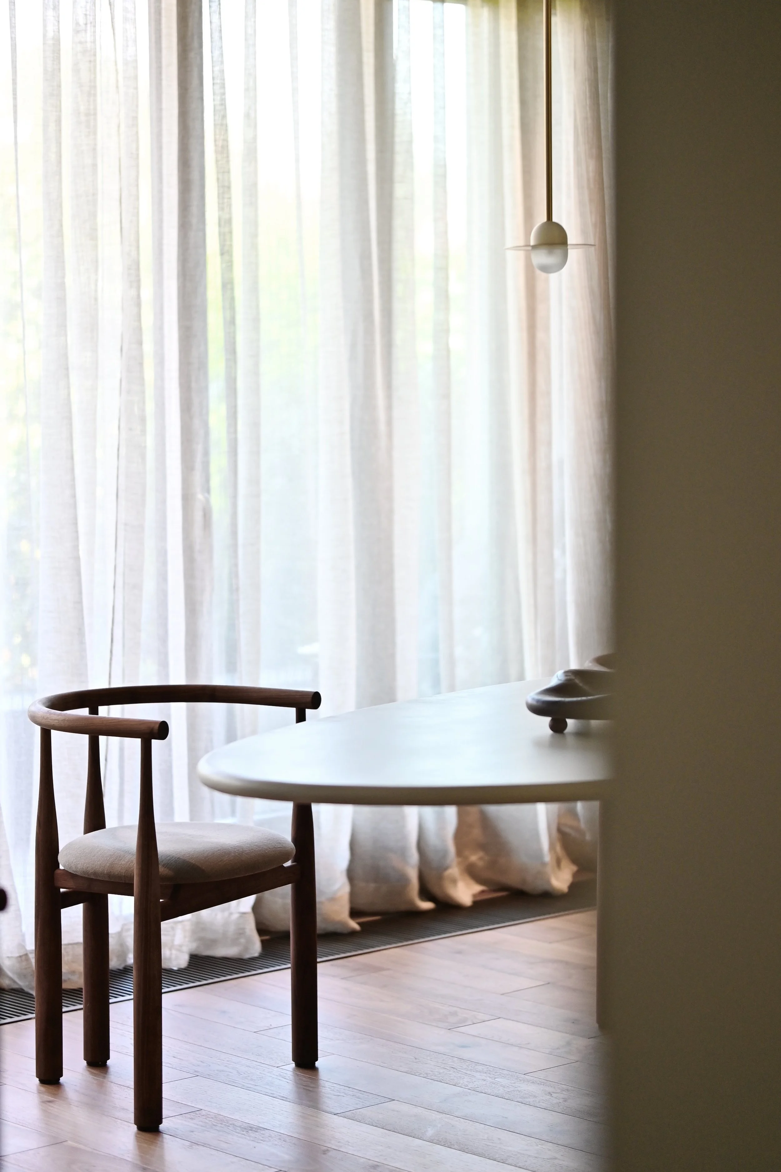 A minimalist interior scene with a wooden chair, a white round table, sheer curtains letting in natural light, a modern pendant lamp, and a portion of a sculpture or decorative object on the table.