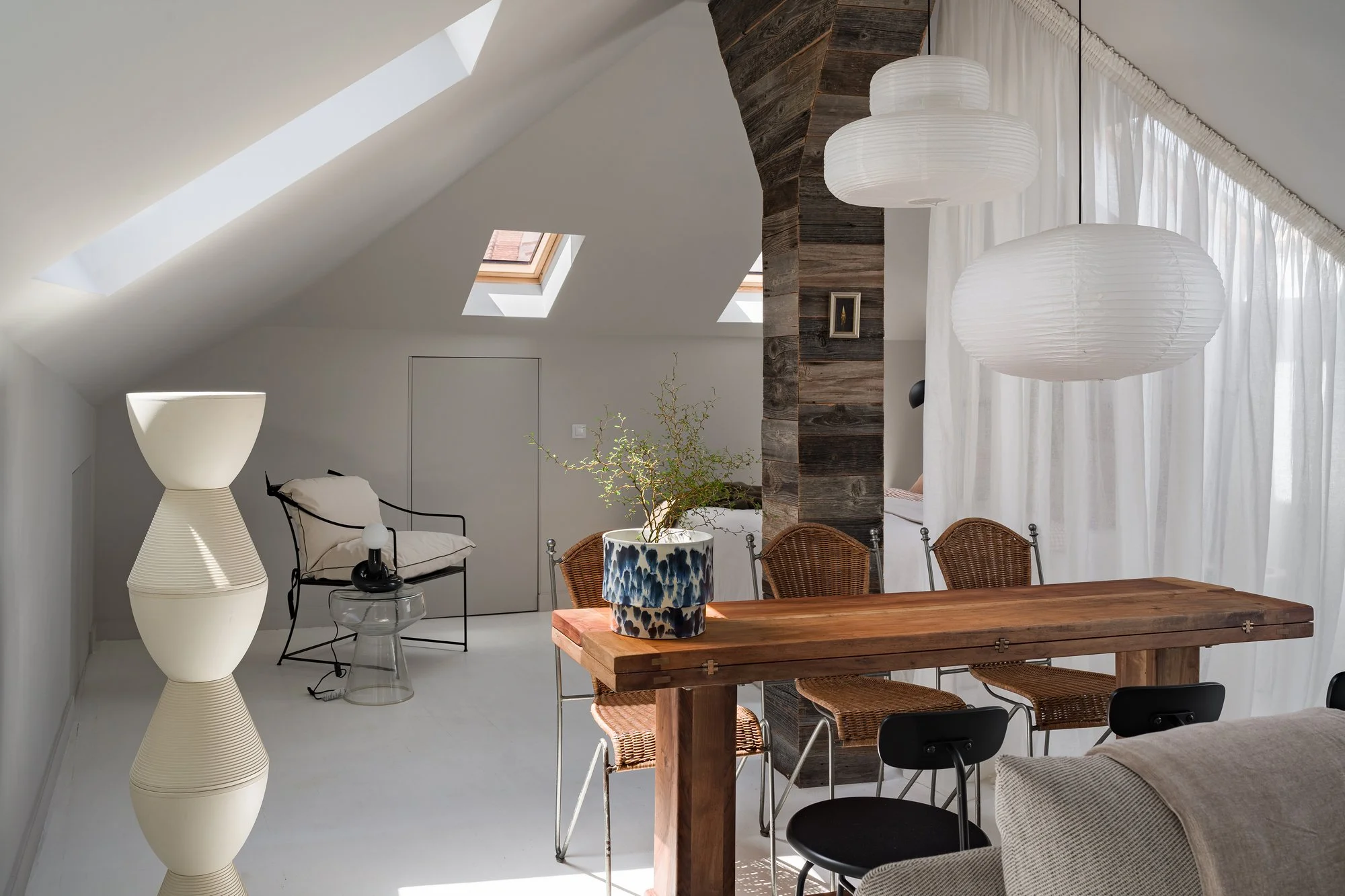 Bright attic room with skylights, white walls, and minimalist decor. Features a wooden dining table with wicker and black chairs, a white sculptural floor lamp, a small side table with a lamp, a potted plant, and sheer white curtains.