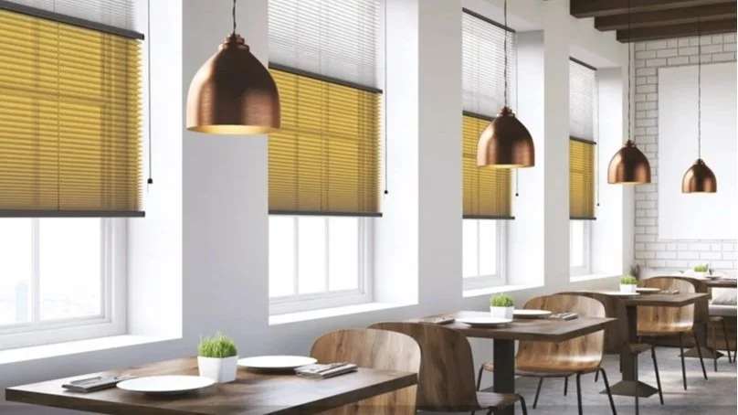 Interior of a modern restaurant with wooden tables, chairs, and ceiling pendant lights. Windows have yellow blinds, and potted plants are on the tables.