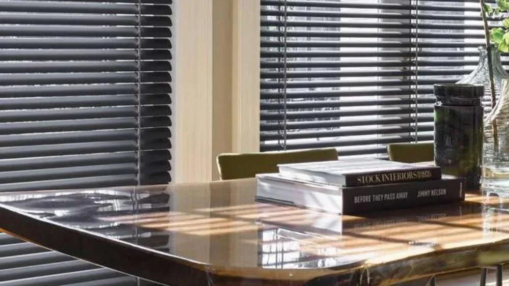 A wooden table with a stack of books and a glass pitcher with greenery, in front of horizontal window blinds.
