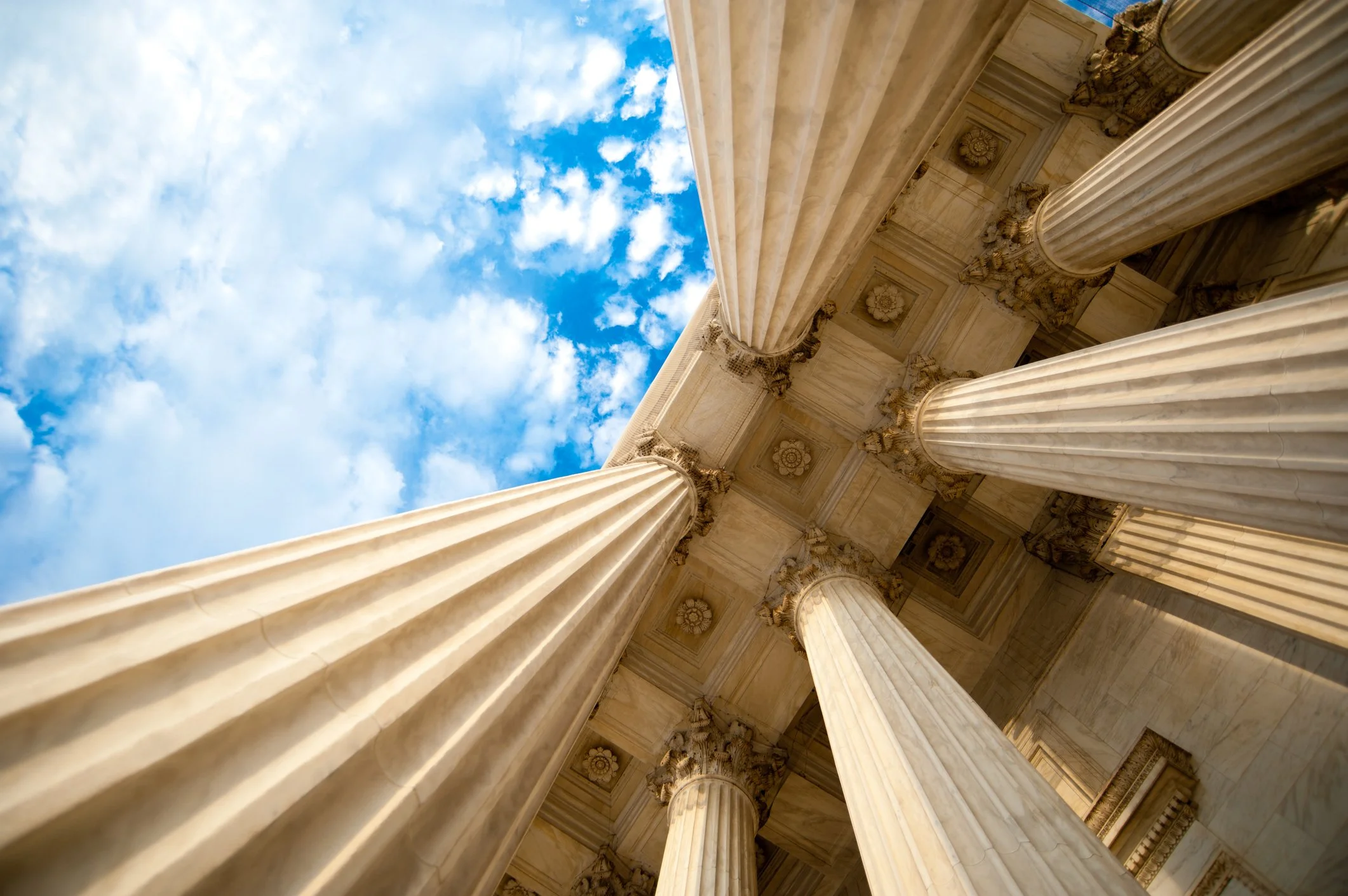 Looking up at grand ancient stone columns and ornate ceiling of a historic building against a bright blue sky with scattered clouds.