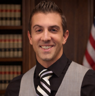 A man smiling in front of bookshelves, wearing a gray vest, black shirt, and striped tie.