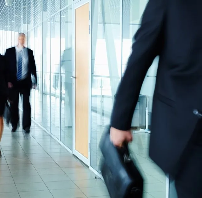 Business professionals walking in a modern office corridor with glass walls, some carrying briefcases.