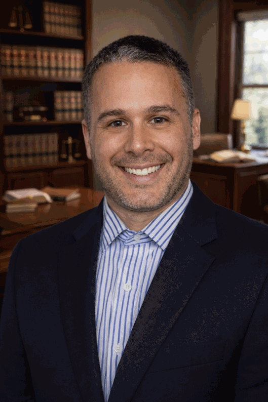 A smiling man in a dark suit and striped shirt standing in a wood-paneled office with bookshelves and a window in the background.