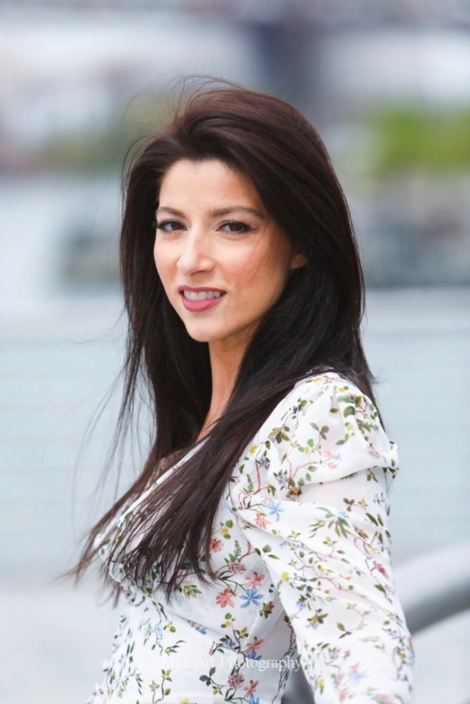 A woman with long dark hair and light skin, smiling slightly, standing outdoors with a blurred urban background.