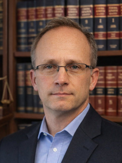A man with glasses and short hair in a suit and blue shirt, standing in front of bookshelves filled with legal books.