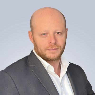 A man with a bald head, light beard, wearing a dark suit jacket and a white dress shirt, posing against a plain light gray background.