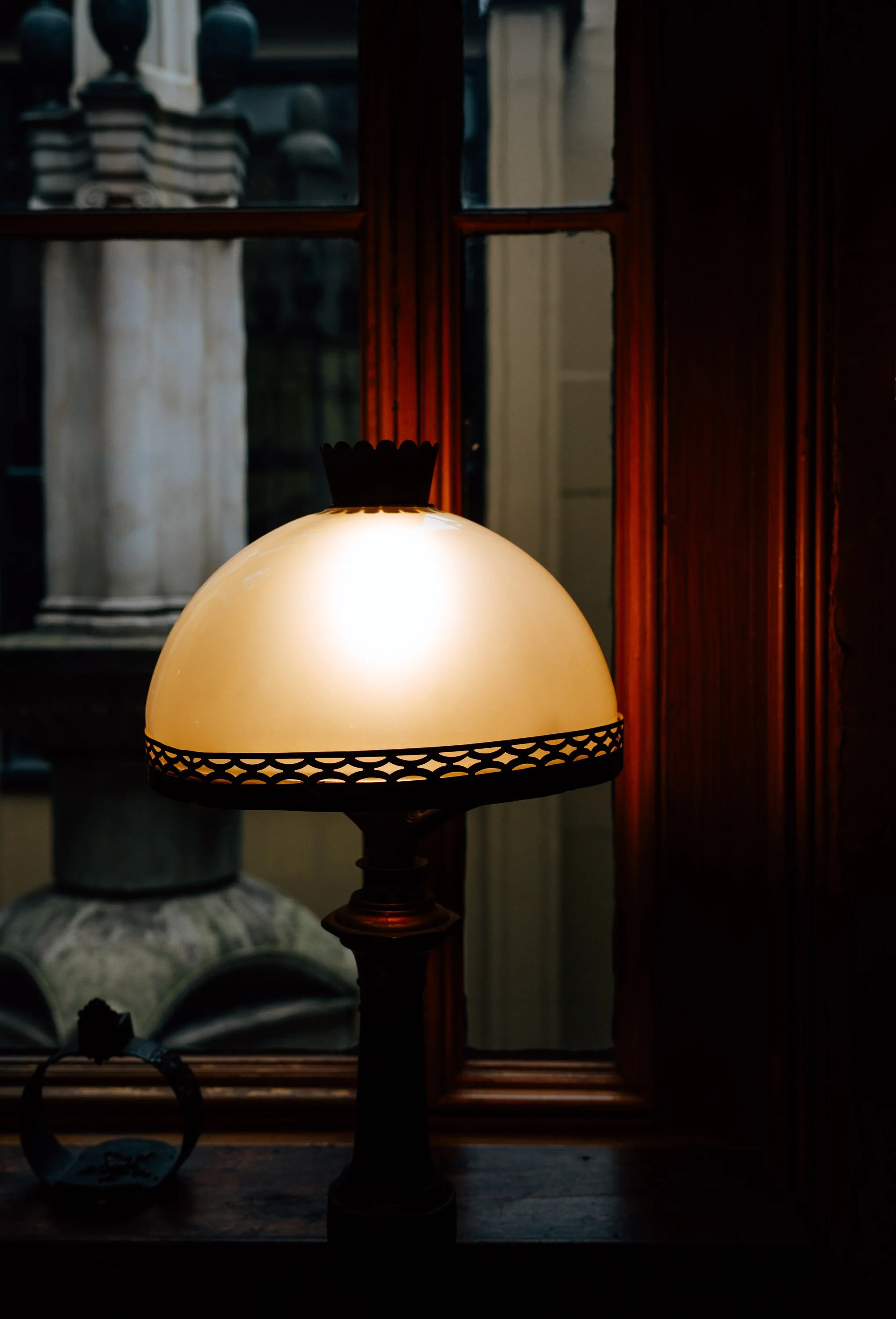 A lit vintage table lamp with a white glass shade and decorative trim, placed on a wooden surface near a window with a wooden frame, showing a view of architectural details outside.