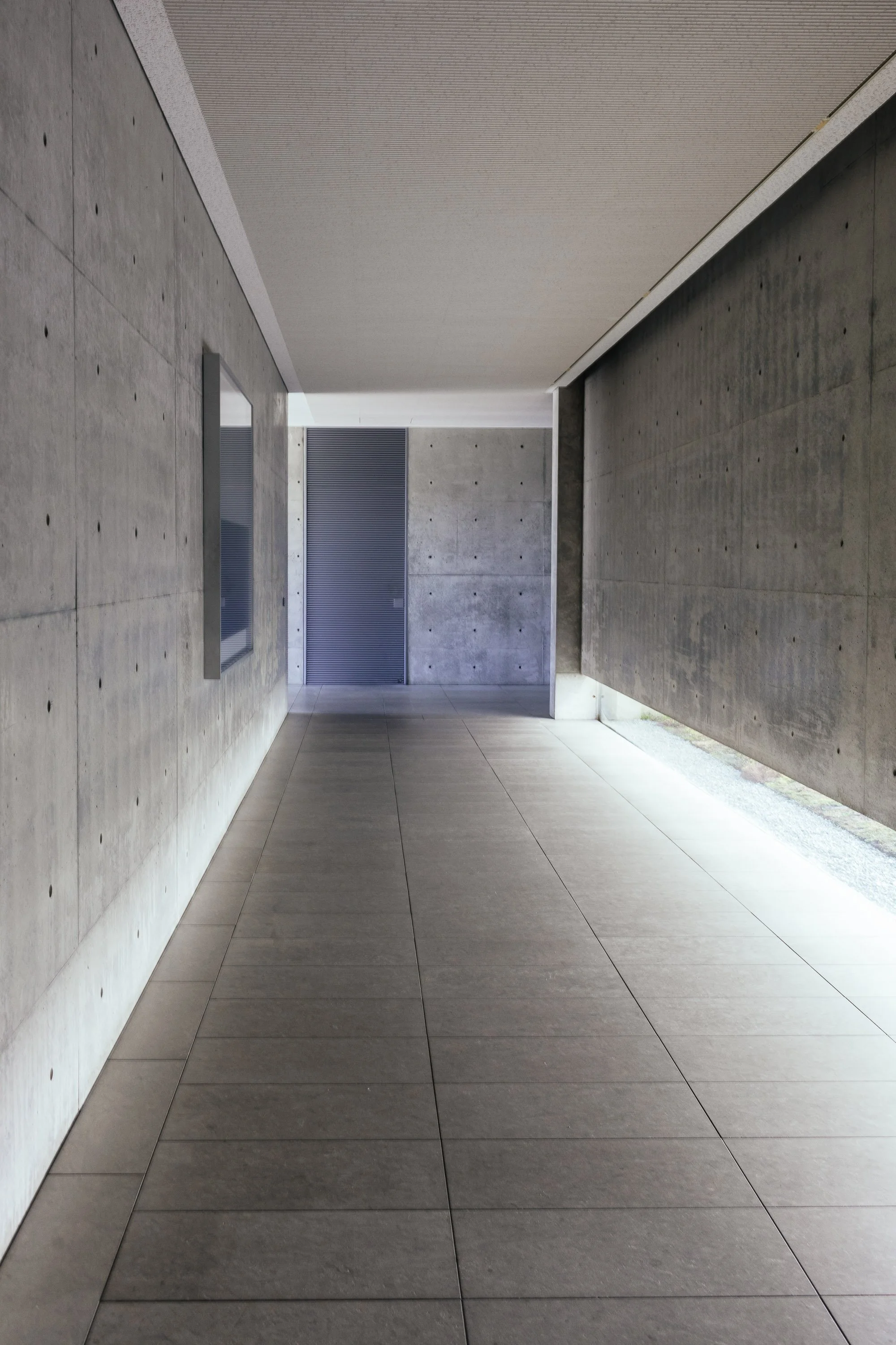 A minimalist, modern interior hallway with concrete walls and tiled flooring, featuring a door with horizontal slats at the end of the corridor.